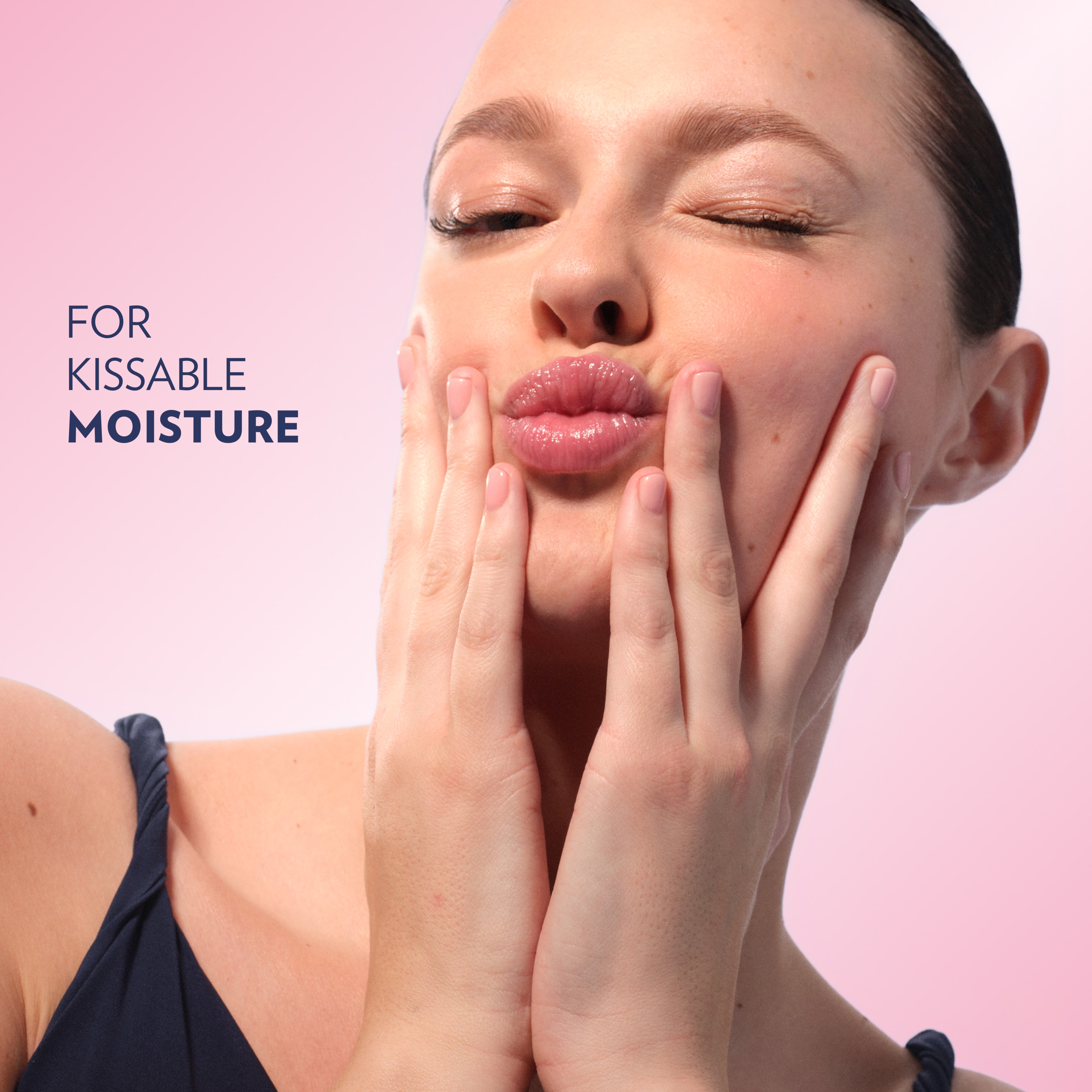 Close-up of a person with glossy lips gently pressing both hands to their cheeks against a pink background, with the text