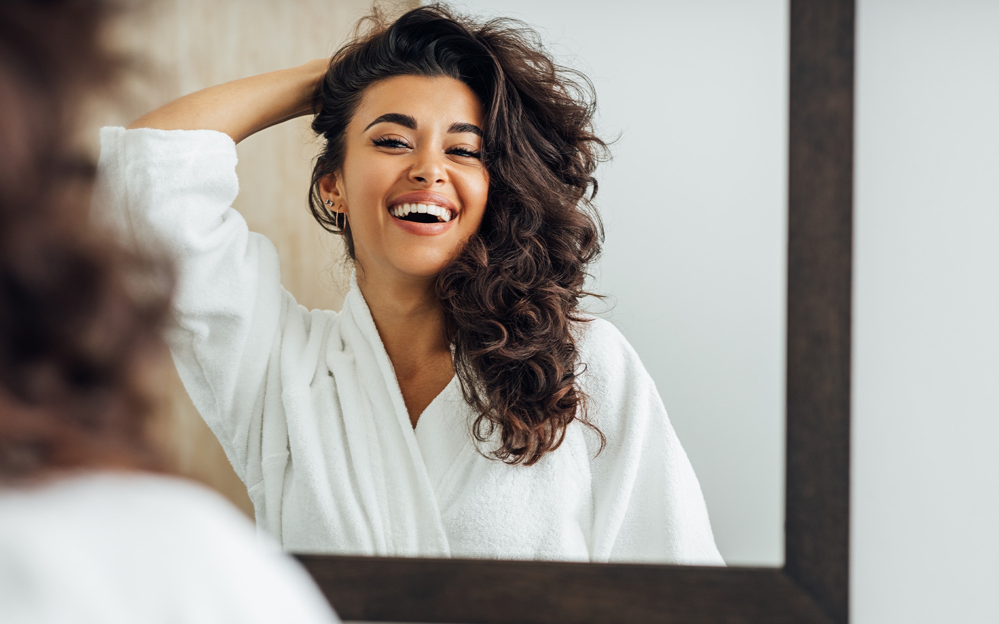 Woman with brown curly hair in a dressing gown smiling into a mirror
