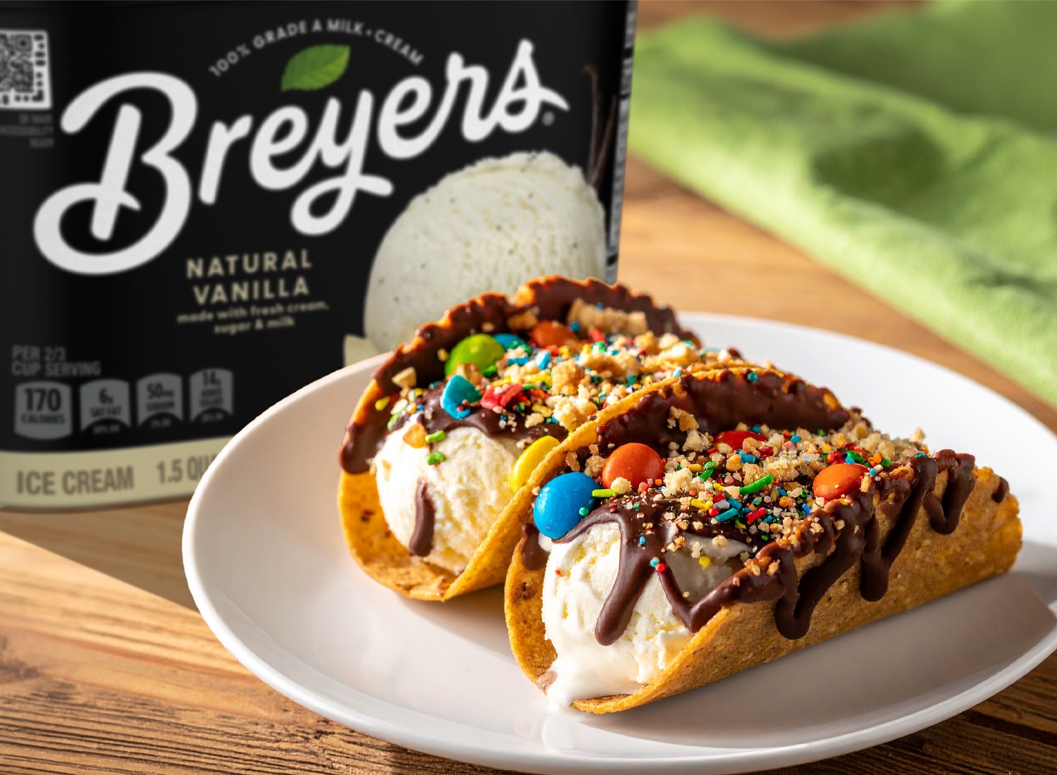 Two ice cream tacos with chocolate drizzle and candy toppings on a plate next to a tub of Breyers Natural Vanilla.