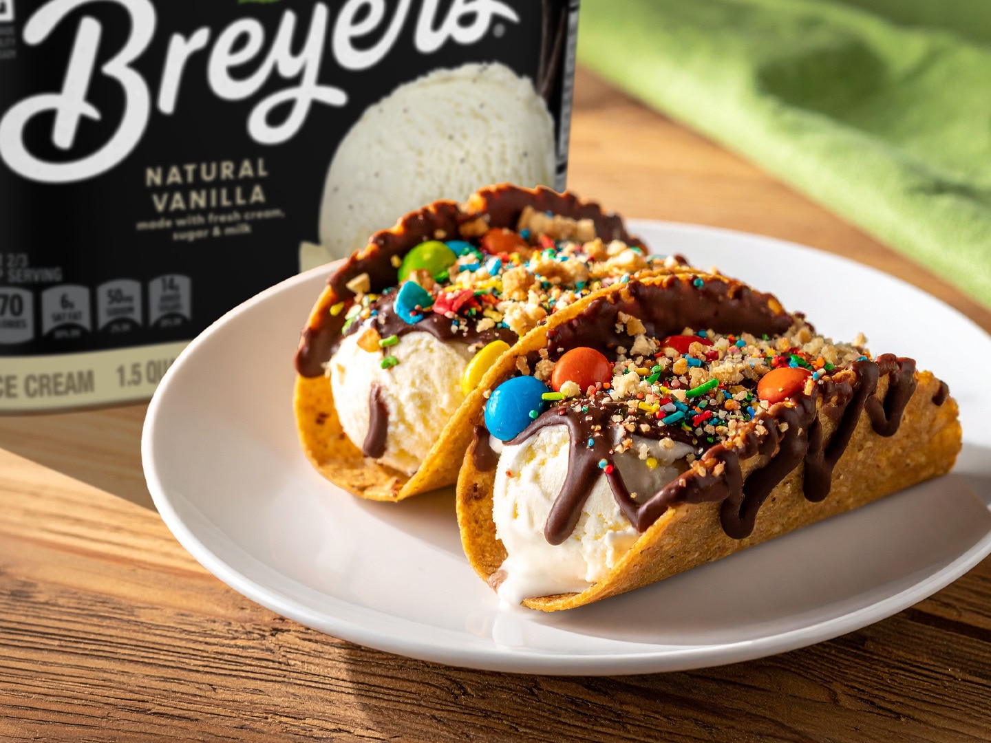 Two ice cream tacos with chocolate drizzle and candy toppings on a plate next to a tub of Breyers Natural Vanilla.