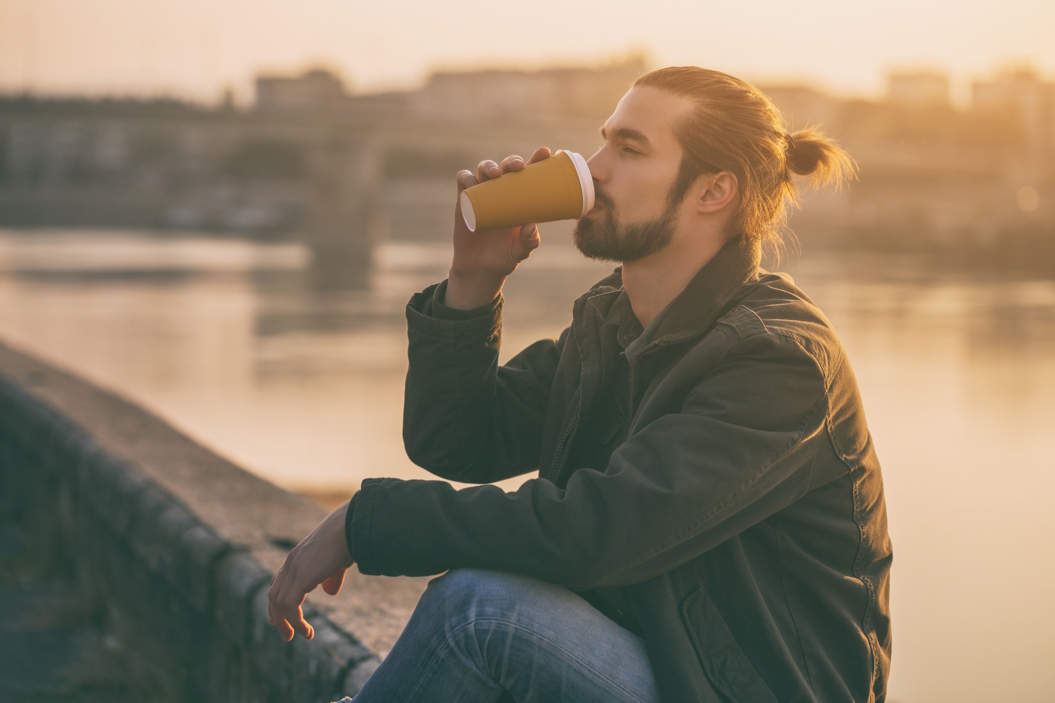 Bearded man with a man bun drinking a takeaway coffee by a river at sunset