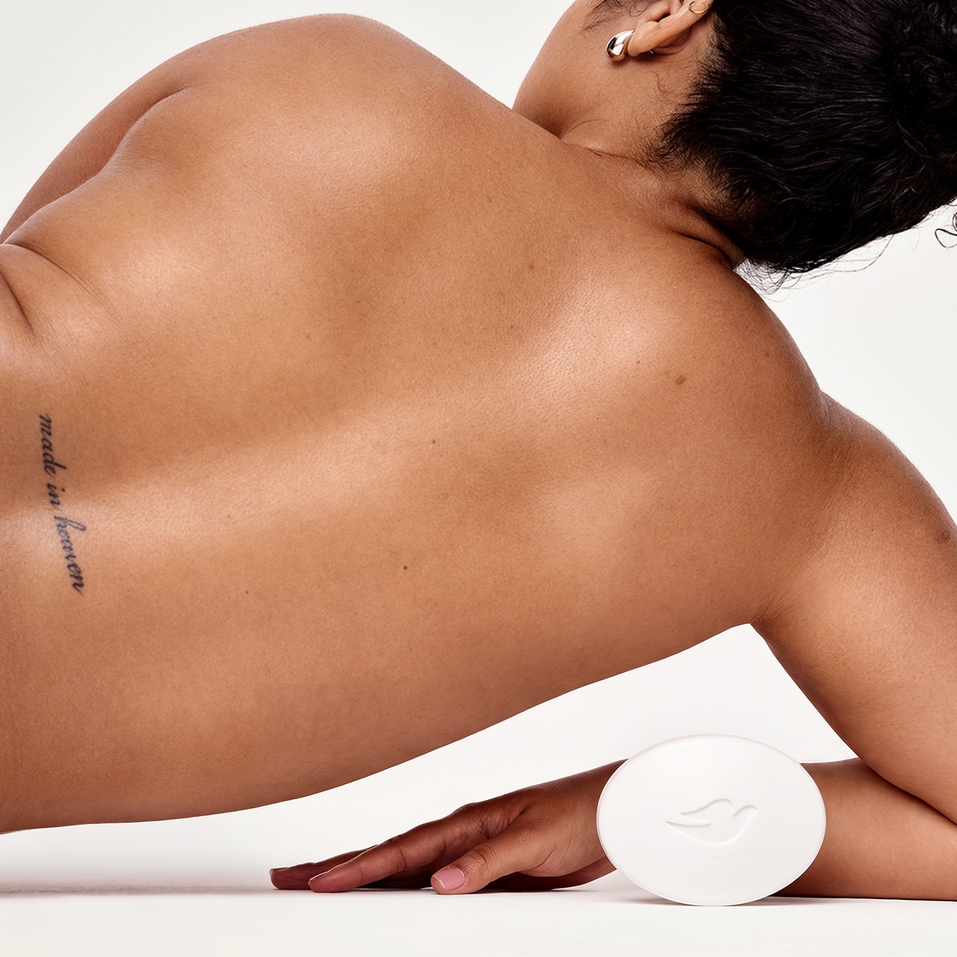 Back view of a woman with a naked back and tattoo on her lower back laying on her side and a white Dove bar balanced next to the back of her wrist.