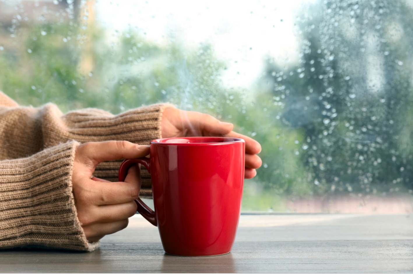 Woman in a beige knitted sweater holding a hot mug on a wooden table