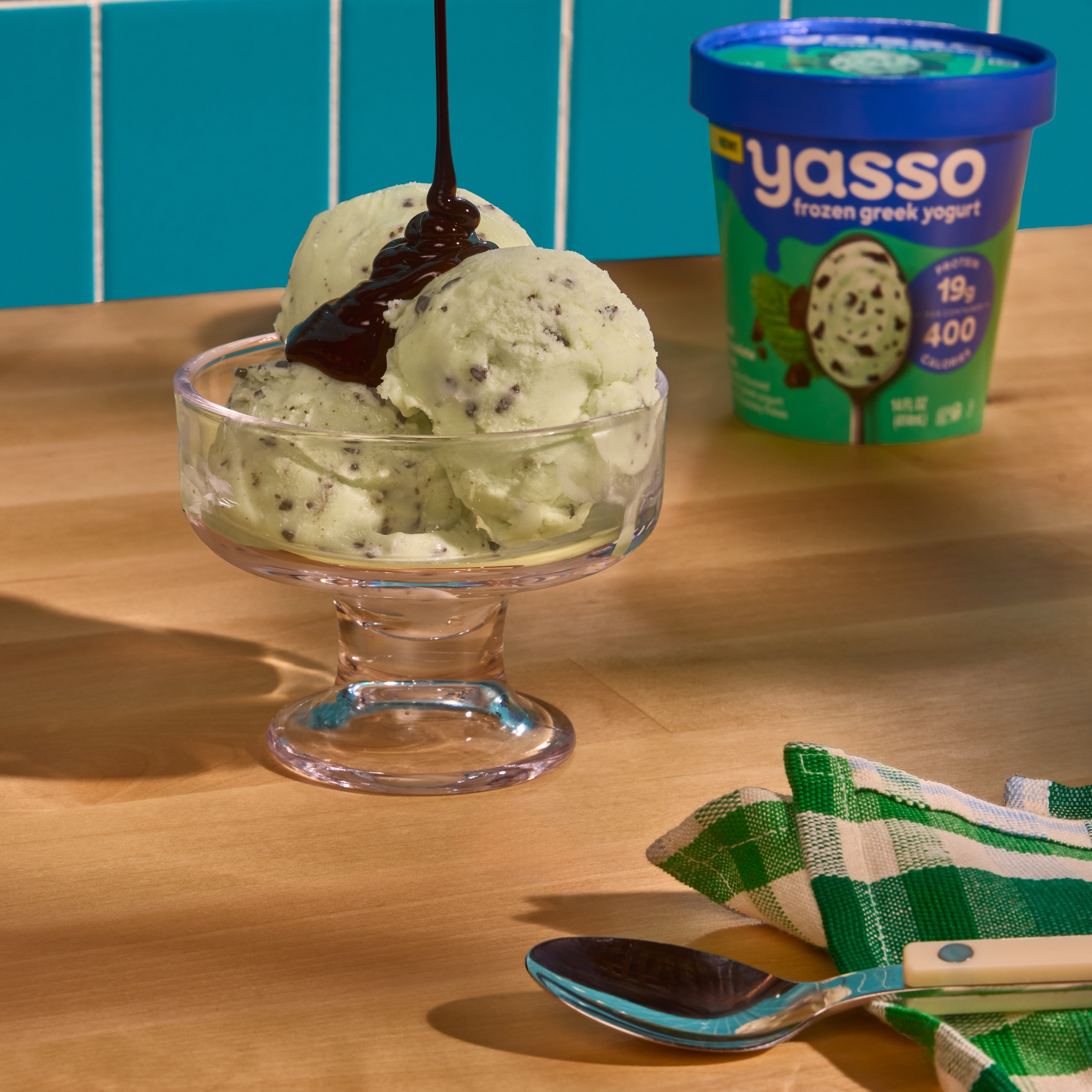 Scoops of Mint Chocolate Chip sit in sundae cup, Pint edges in background. Fudge drizzles from top of frame into cup.