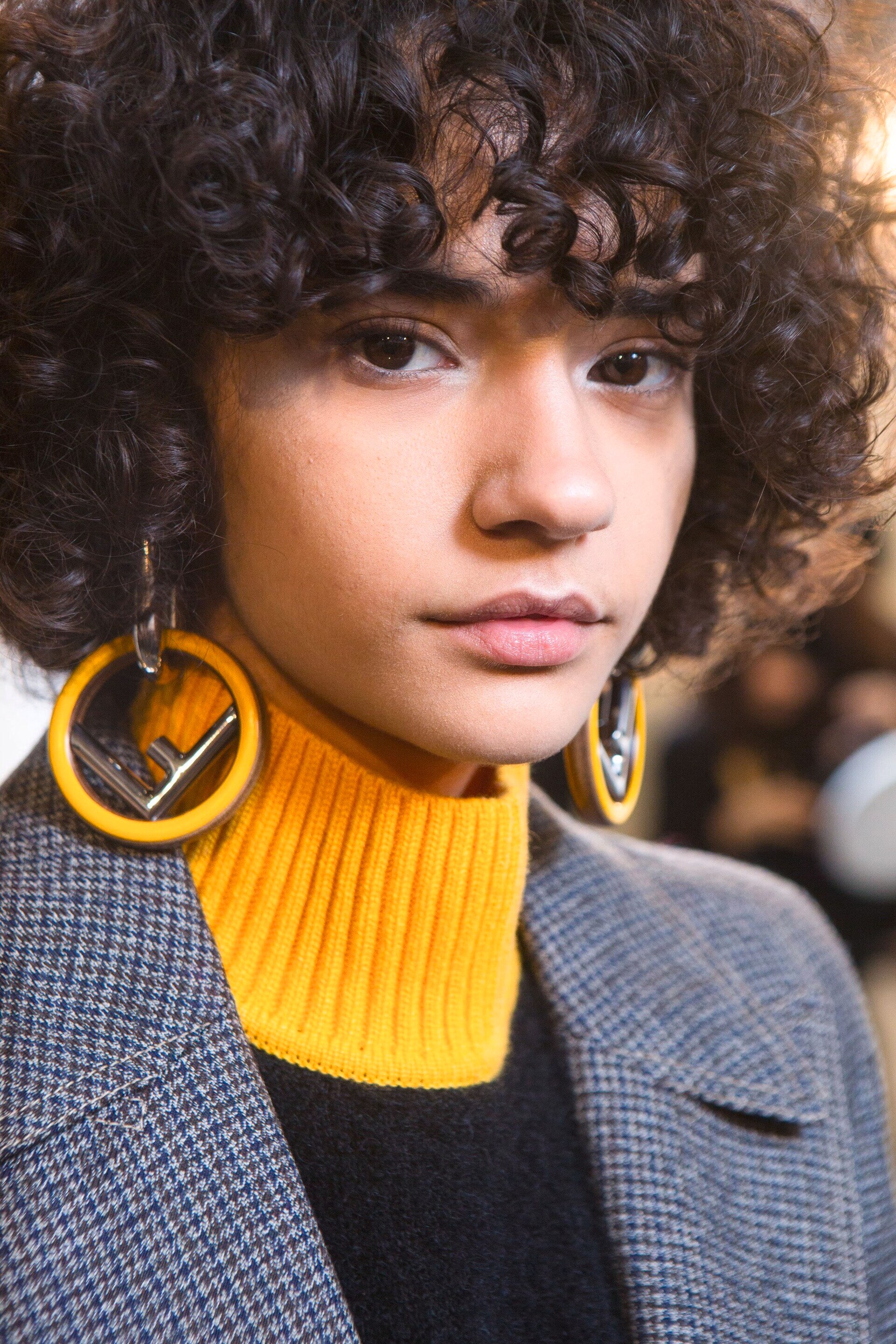 Grown out bangs: Woman with natural brown curly hair cut into a bob and bangs wearing a grey blazer and yellow turtle neck sweater.