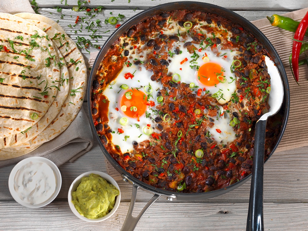huevos rancheros served in frying pan with a spoon, placed on a wooden surface. 