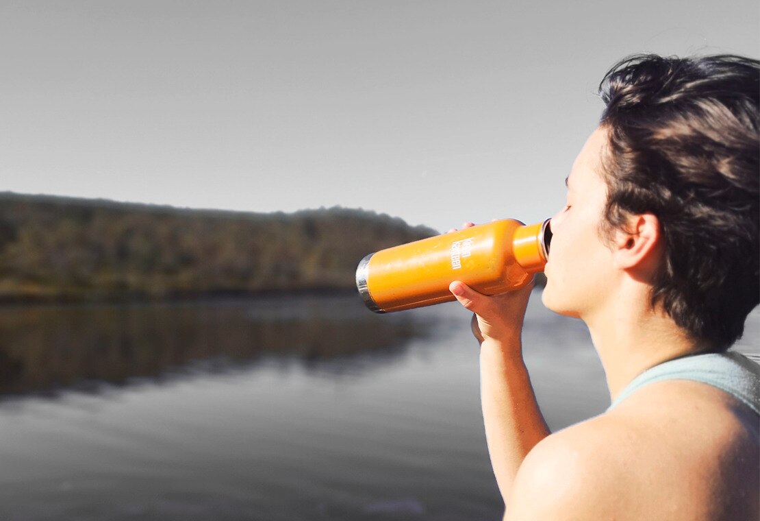 woman by lake drinking from an orange water bottle
