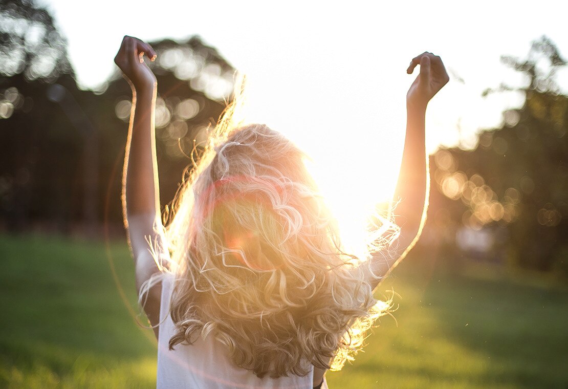 Woman with curly blonde hair raising her arms as the sun sets