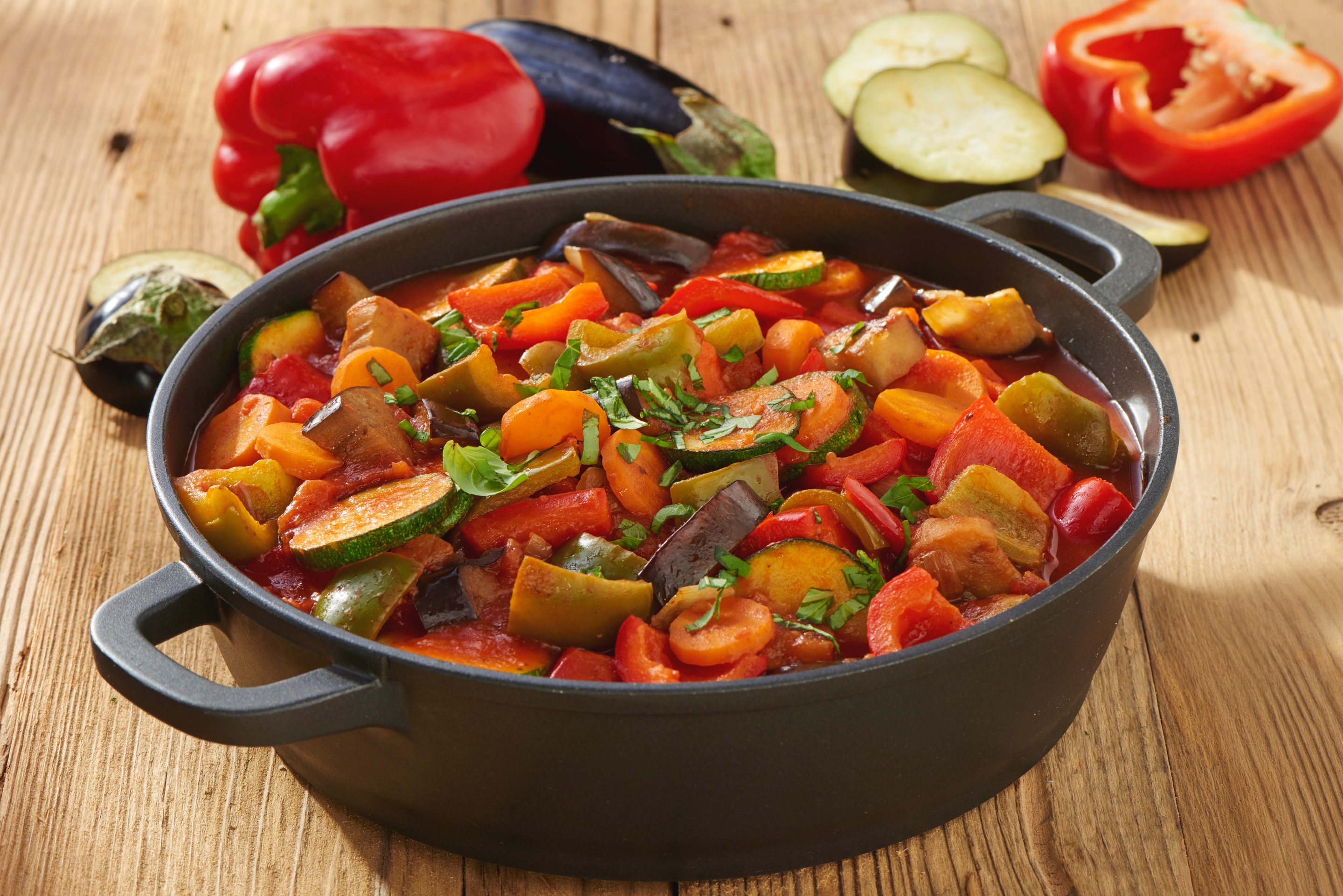 A close shot of colorful ratatouille served in casserole kept on wooden table. Red capsicum and brinjal kept next to casserole.