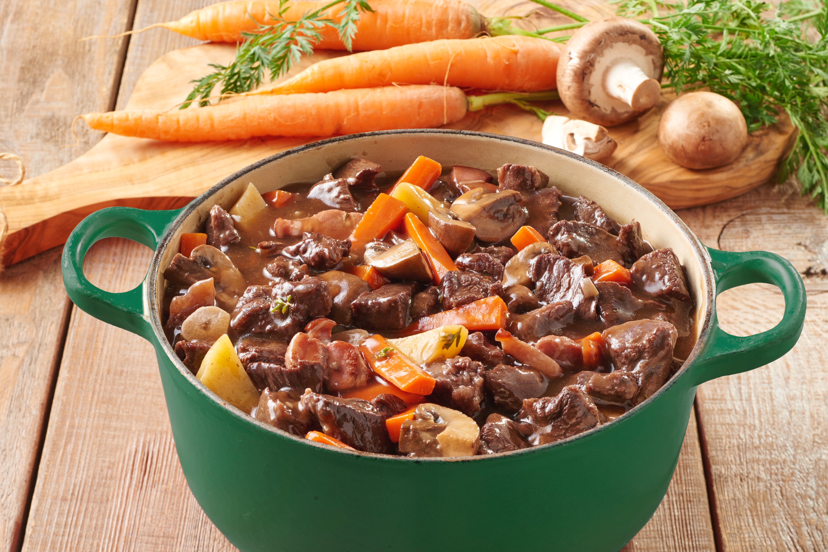 Rich Beef Bourguignon containing carrots, potatoes and mushrooms in an orange casserole pot.  In the background there is carrots and mushrooms on a wooden chopping board.