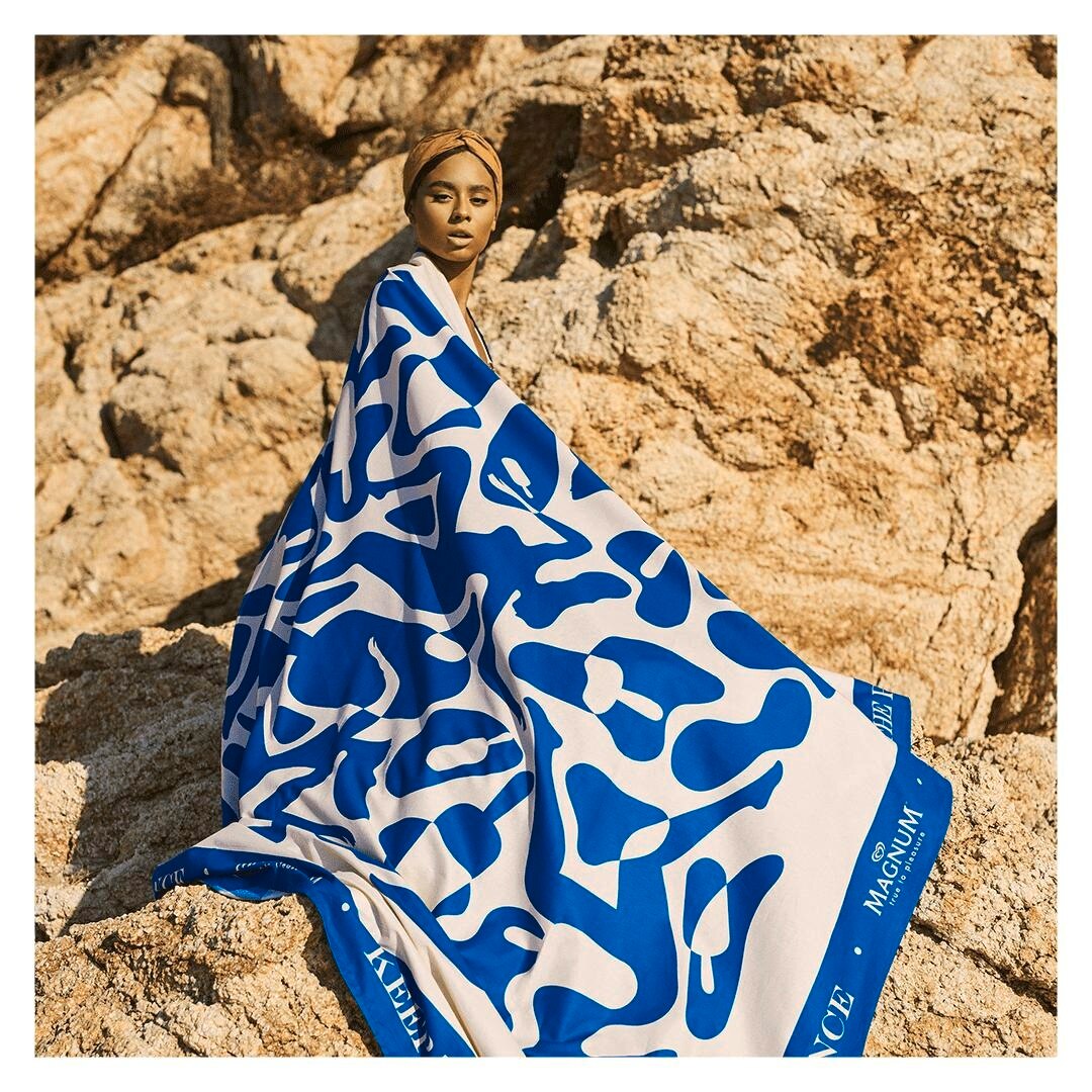 Mulher com uma bandana no cabelo sentada em um rochedo com o corpo inteiro coberto pela toalha gigante Text