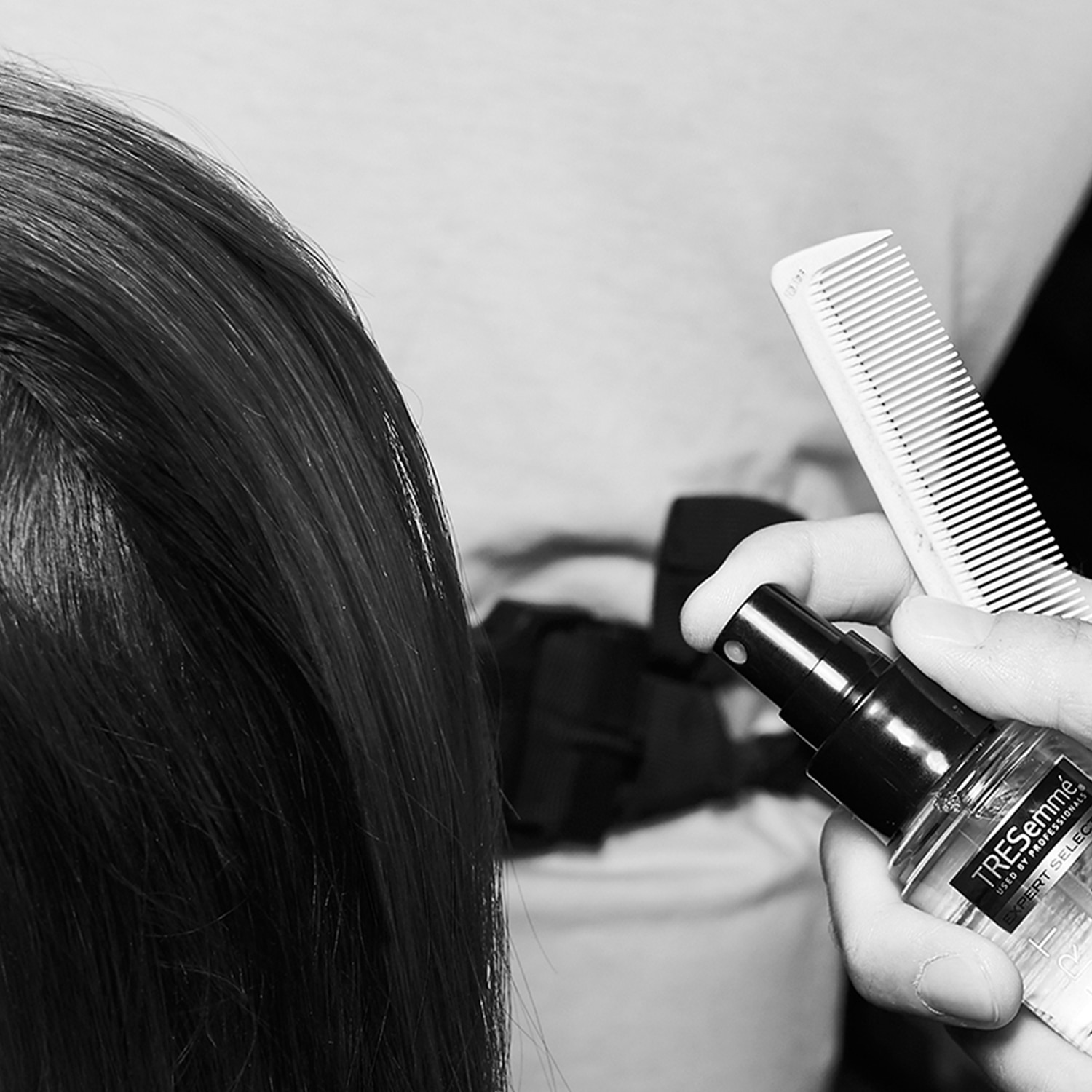 Black and white close up shot of the side of a woman's head with a parting in her hair, plus the hands of a stylist holding a comb and TRESemmé hair care product