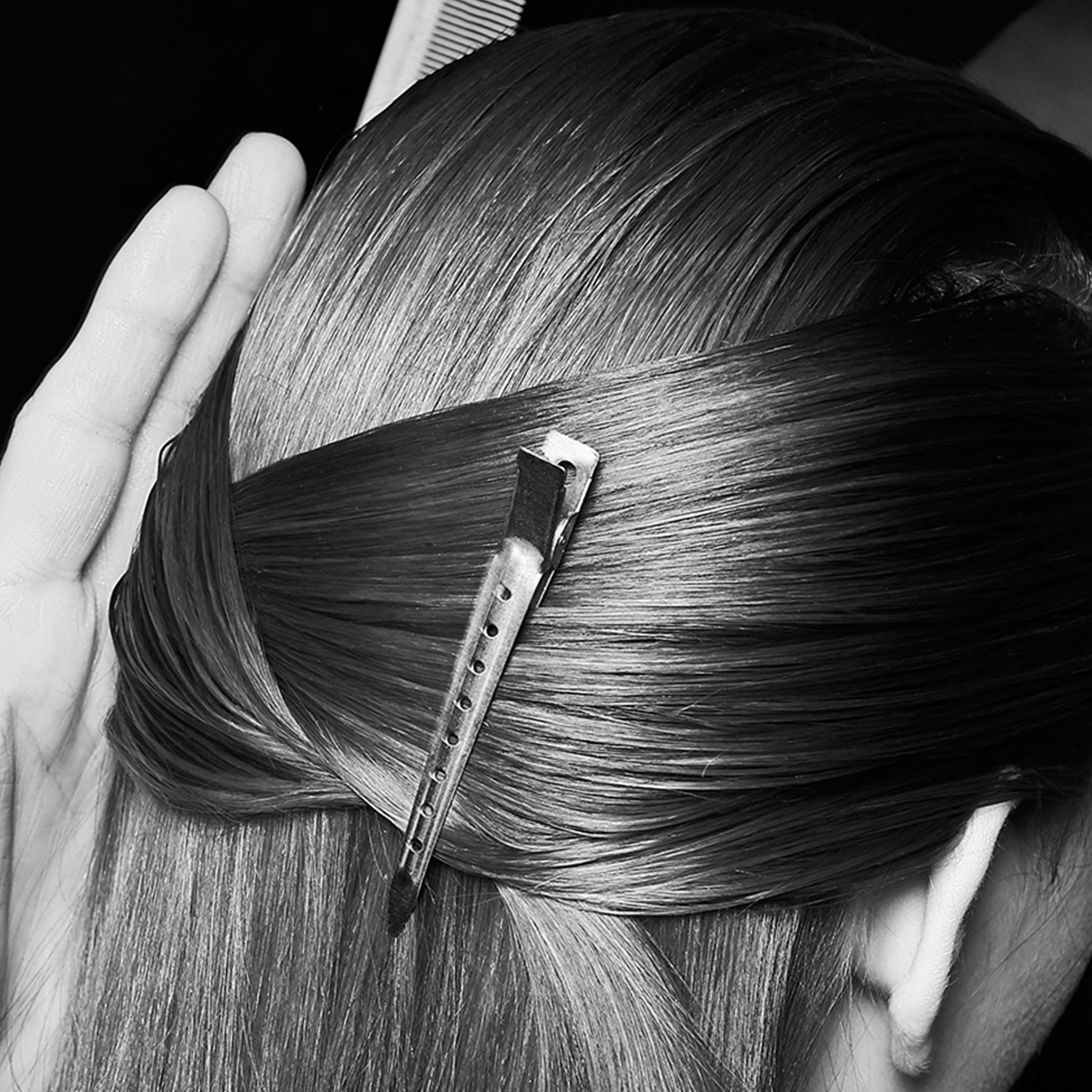 Black and white close up shot of the back of a model's head with the sides of her hair gathered in a clip at the back, with a stylist's hands and comb just visible 