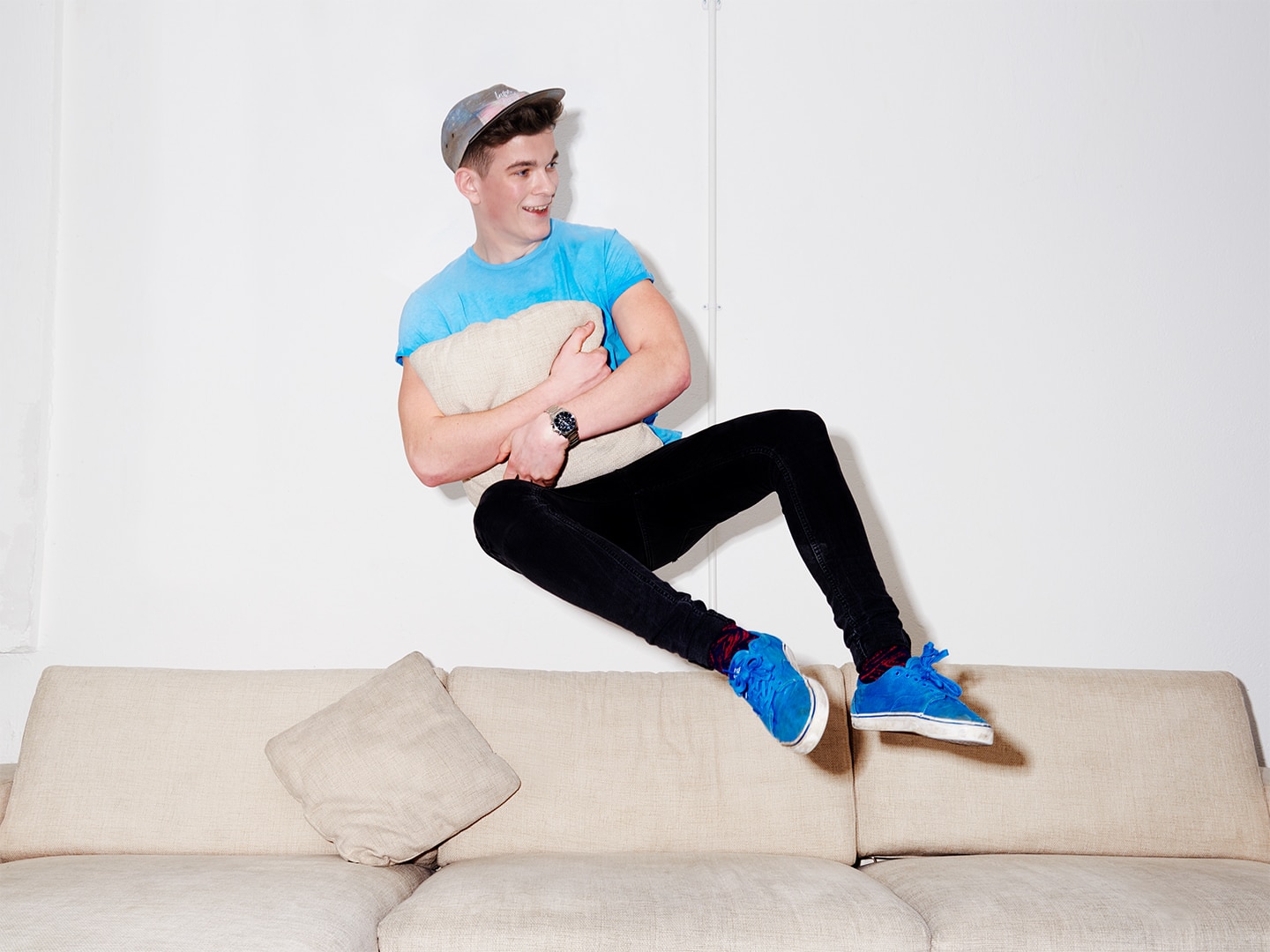 Guy in blue t-shirt and cap falling onto a sofa holding a cushion
