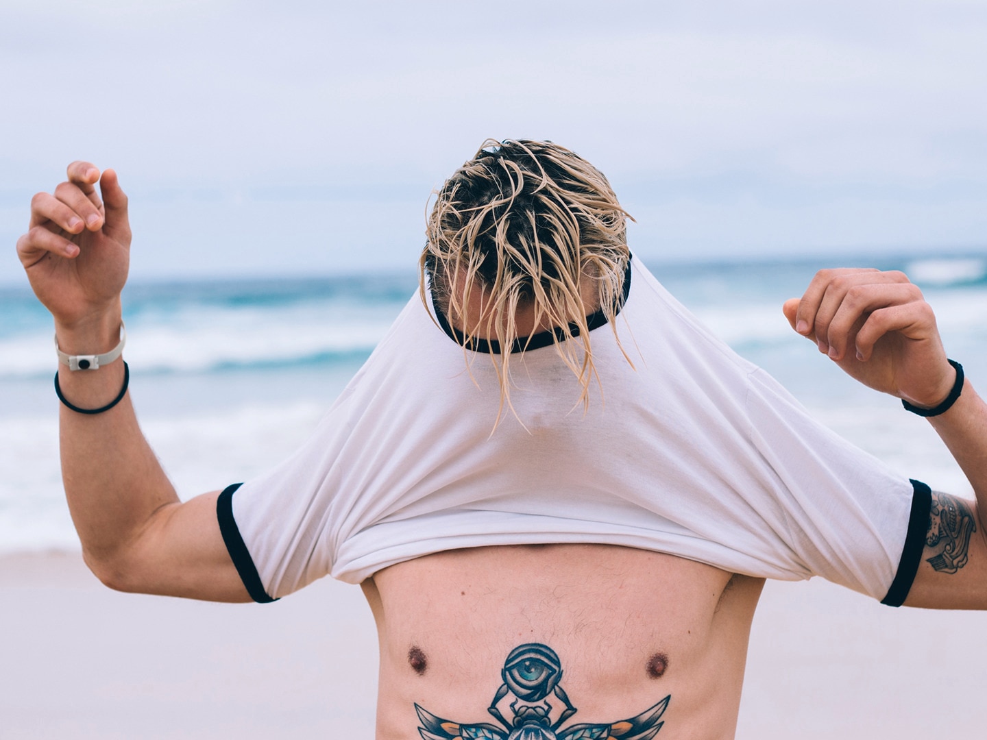 Man with tattoos pulling t-shirt over head on the beach