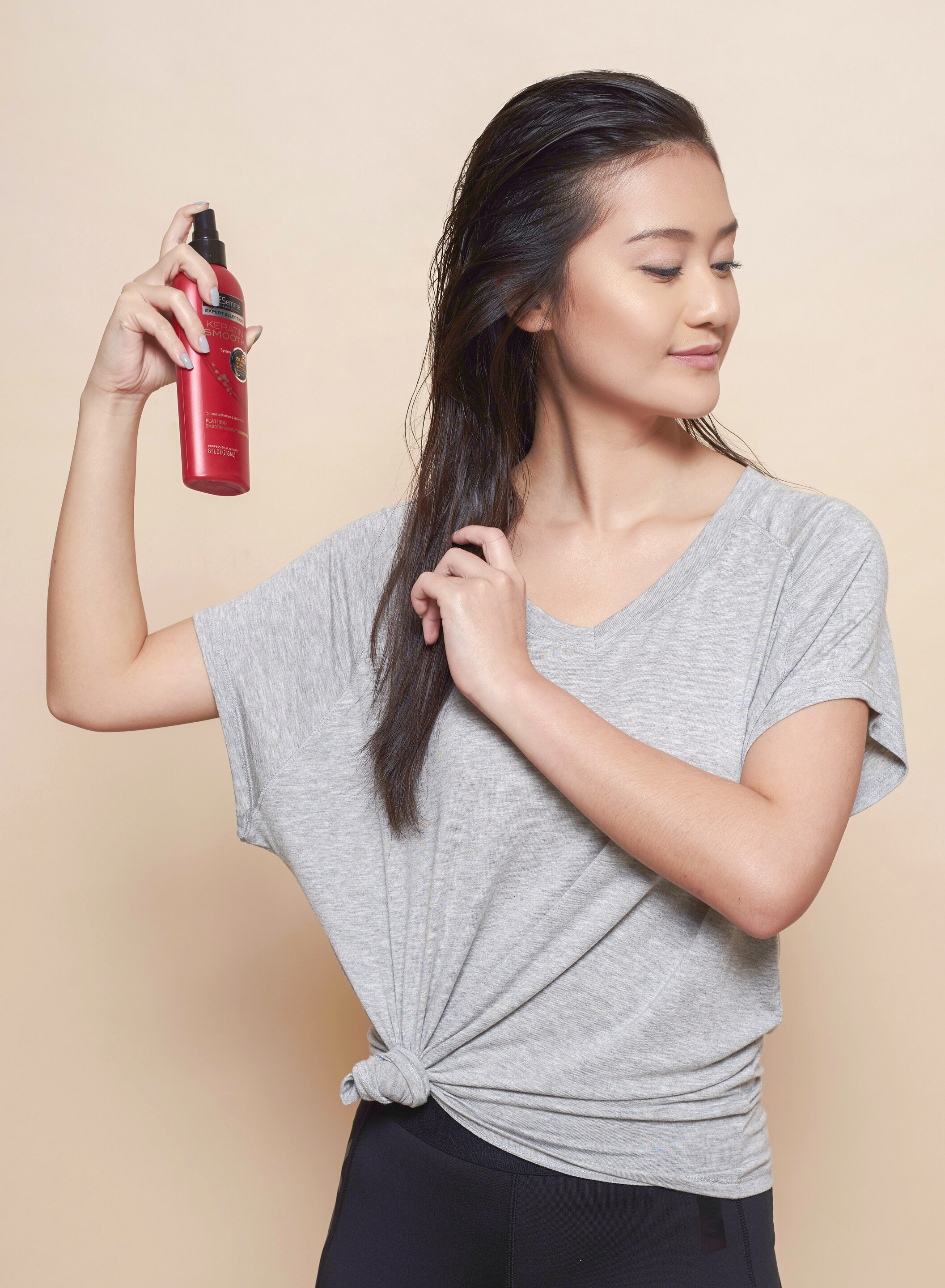 Wanita asia dengan atasan abu-abu dan warna rambut hitam sedang menyemprotkan heat protection spray pada rambut