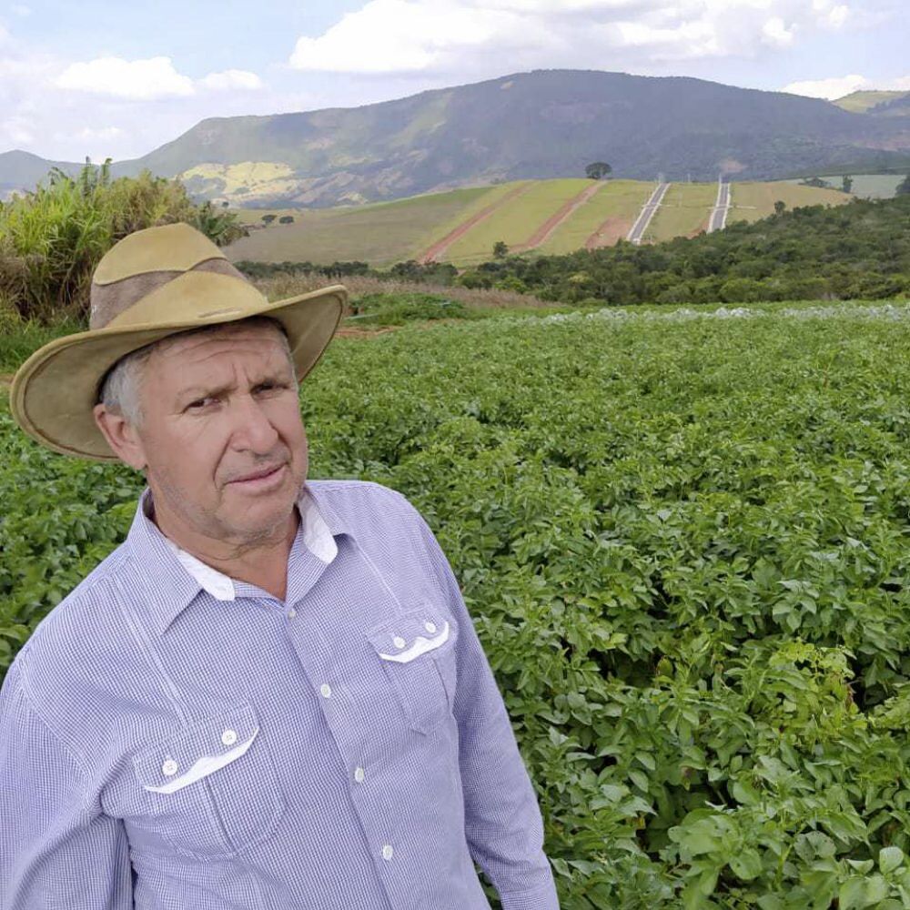 Um idoso de camiseta e chapéu está em pé diante de uma plantação