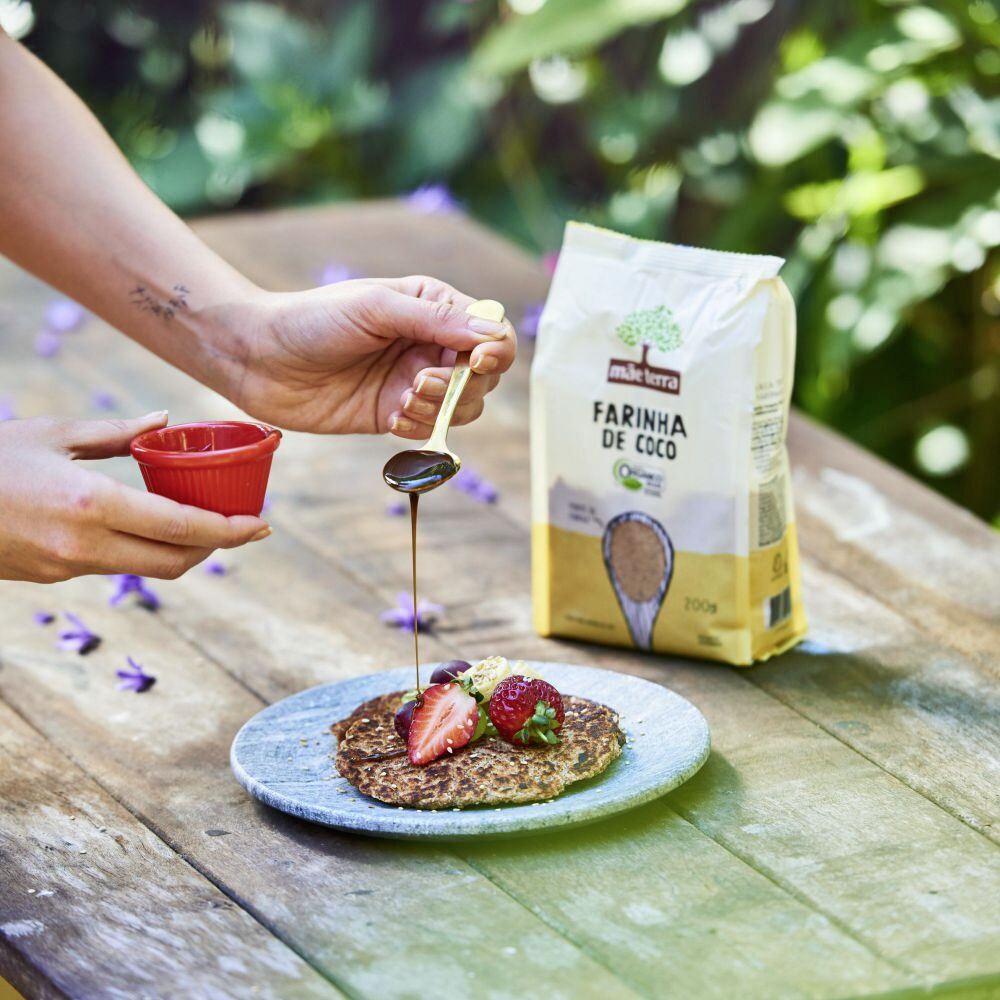 Alguém cobre uma panqueca e morangos com chocolate ao lado da Farinha de Coco Mãe Terra