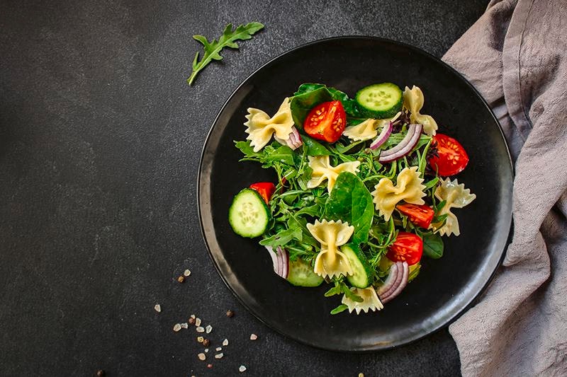 Salad zucchini, tomat, rocket, dan farfalle disajikan dalam mangkuk hitam