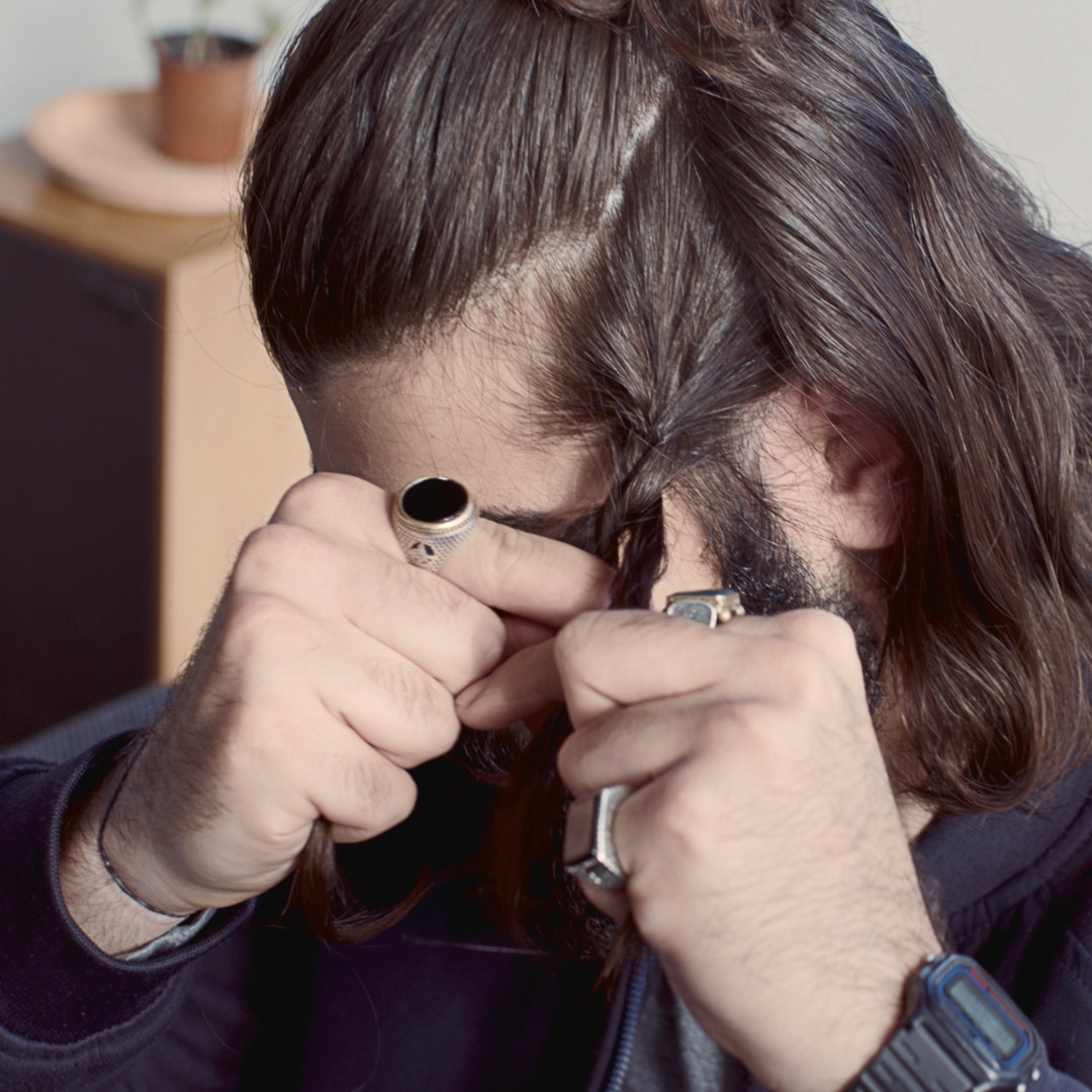 A guy wearing big rings, braiding his hair.