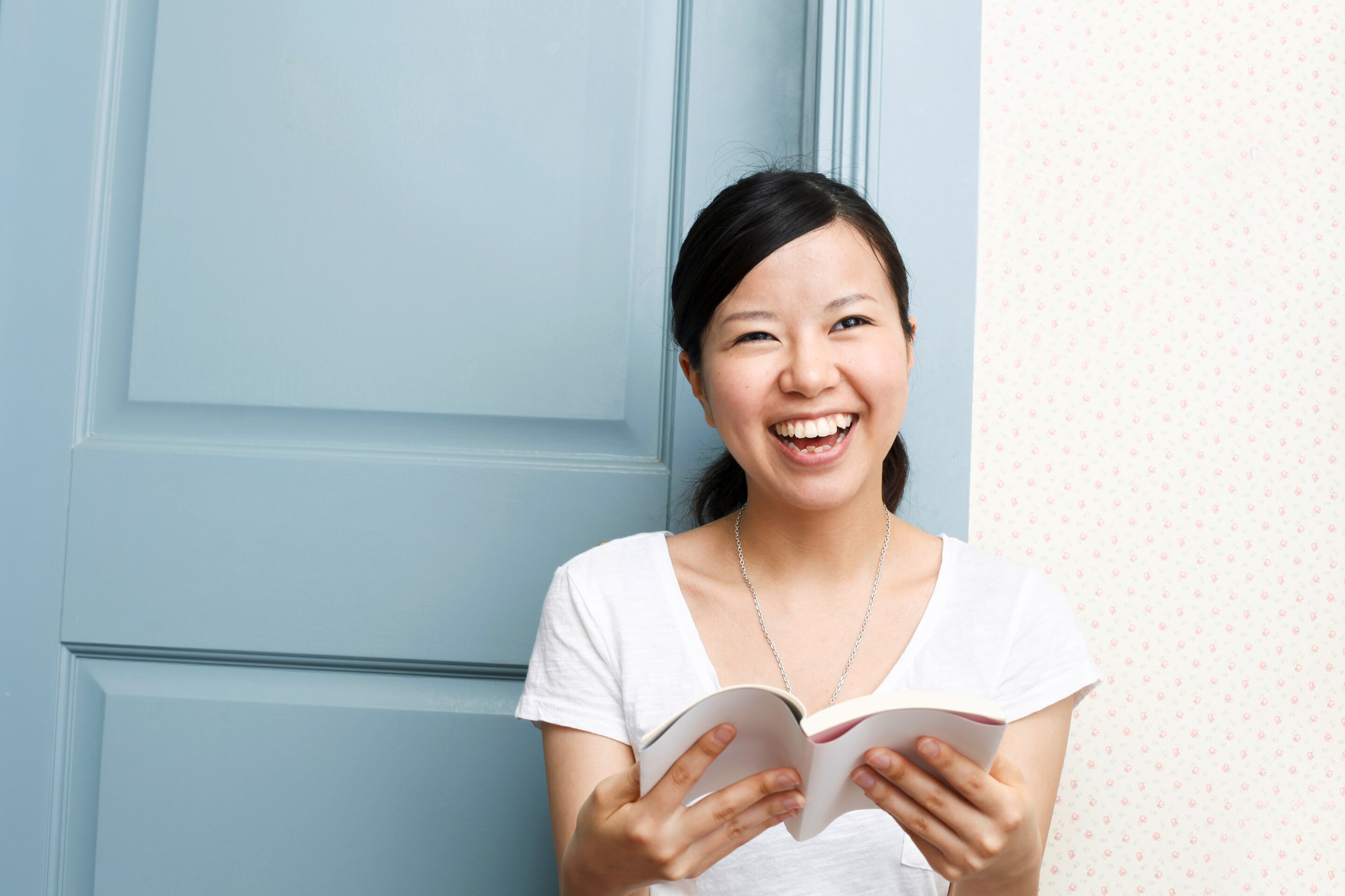 An Asian woman against a blue door, laughing while holding a book