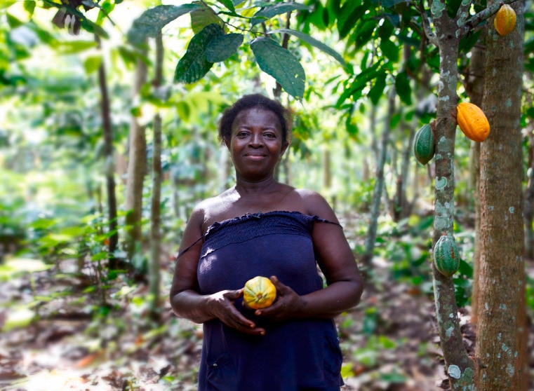 Lächelende Kakaobäuerin, die eine Kakaofrucht hält, umgeben von Bäumen.