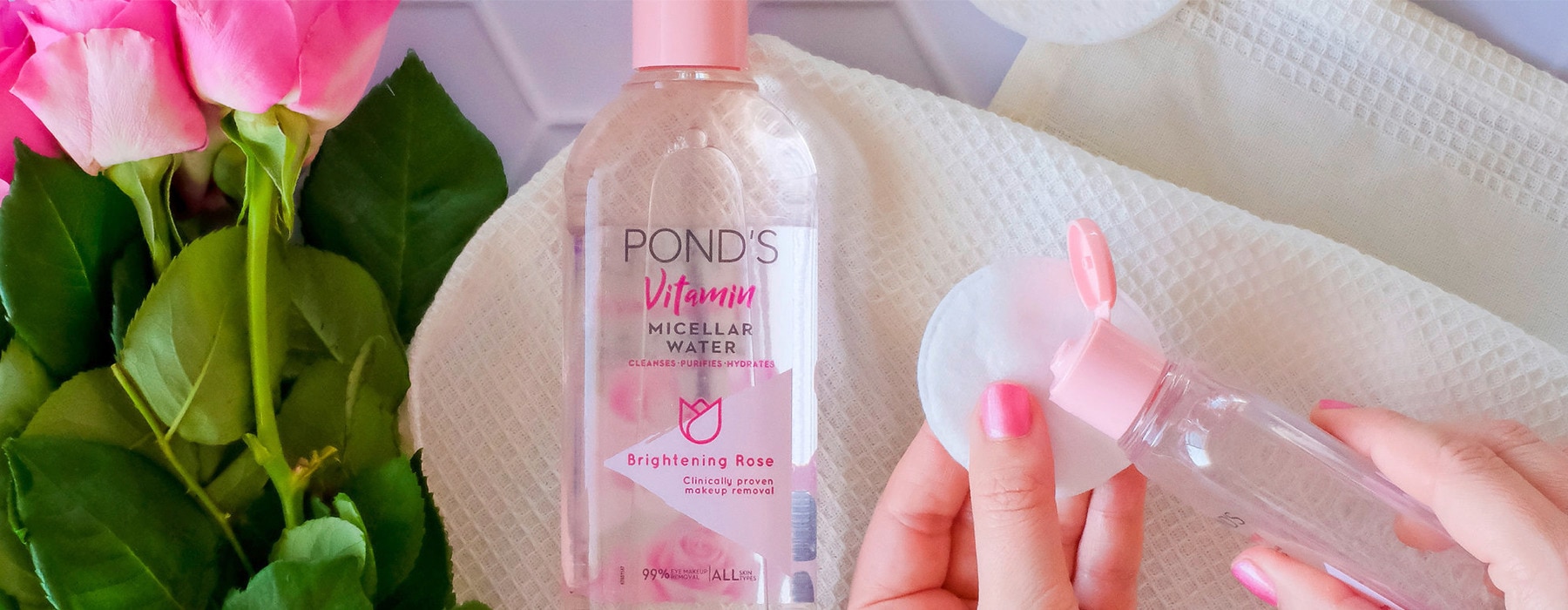 A person pouring micellar water on a makeup remover pad next to the pink roses and make up brush
