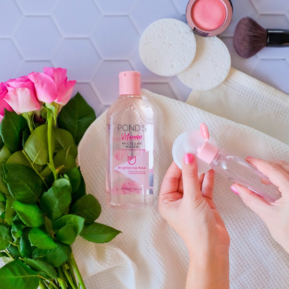 A person pouring micellar water on a makeup remover pad next to the pink roses and make up brush