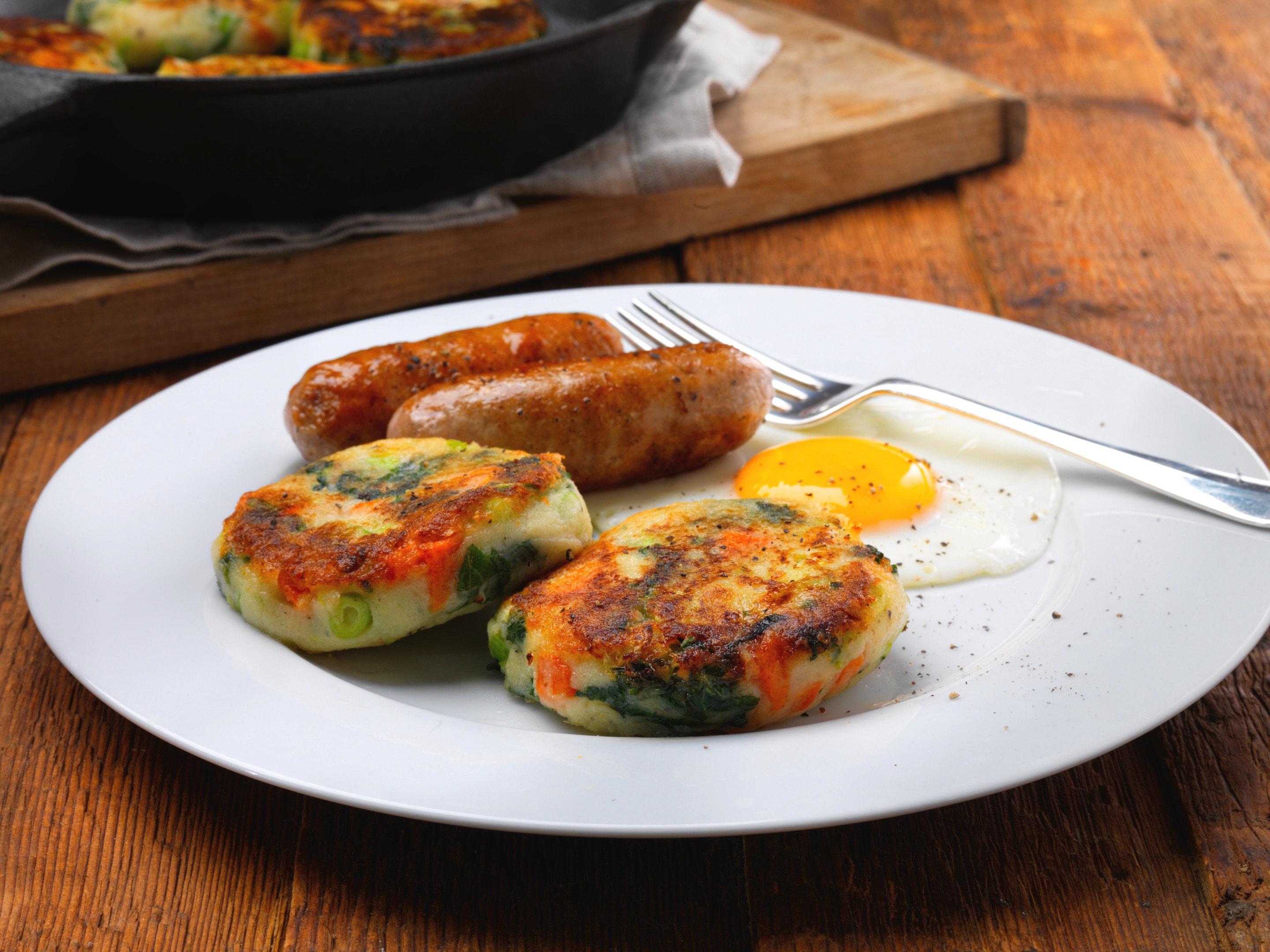 A plate of Bubble and squeak which is a traditional English dish made with the shallow-fried leftover vegetables, grilled sausages and a half cooked egg is placed in a dish on a wooden surface.