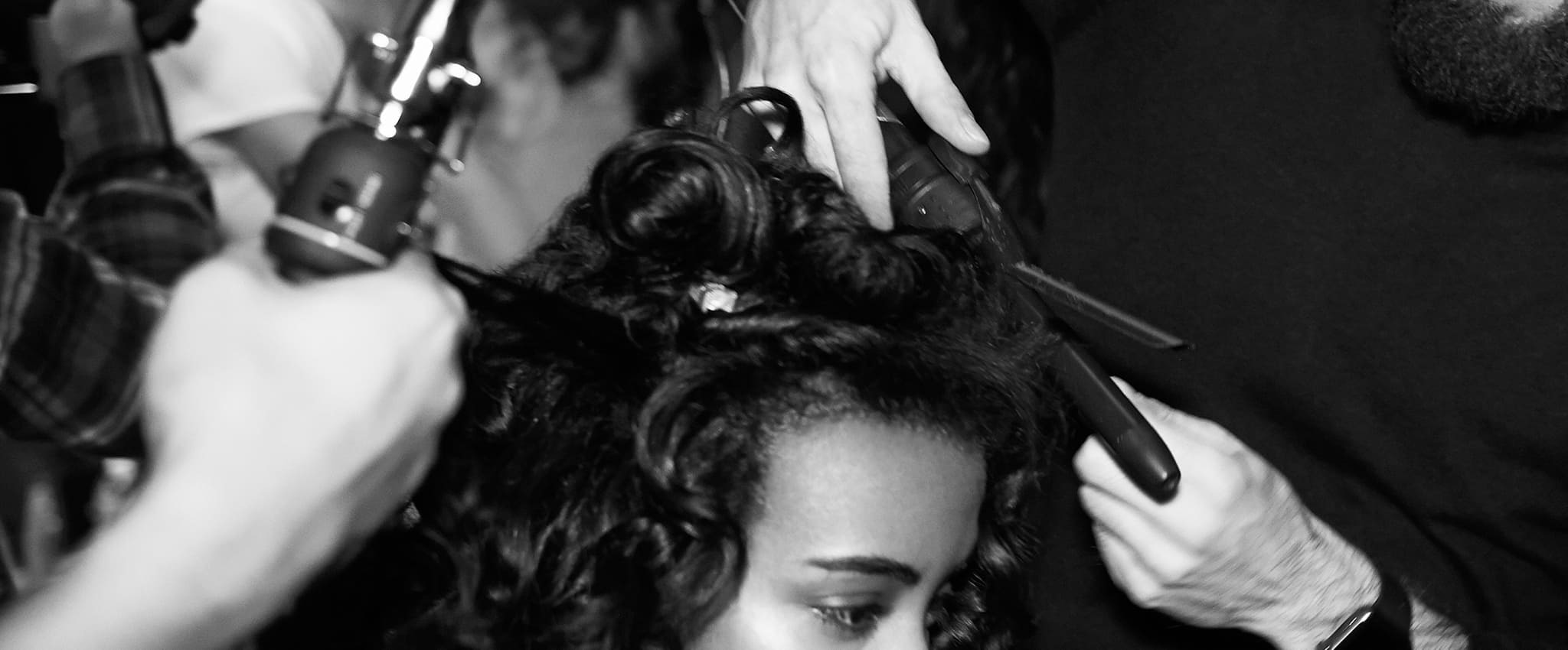 A model has her curly hair worked on by two stylists using heat tongs and other styling tools.