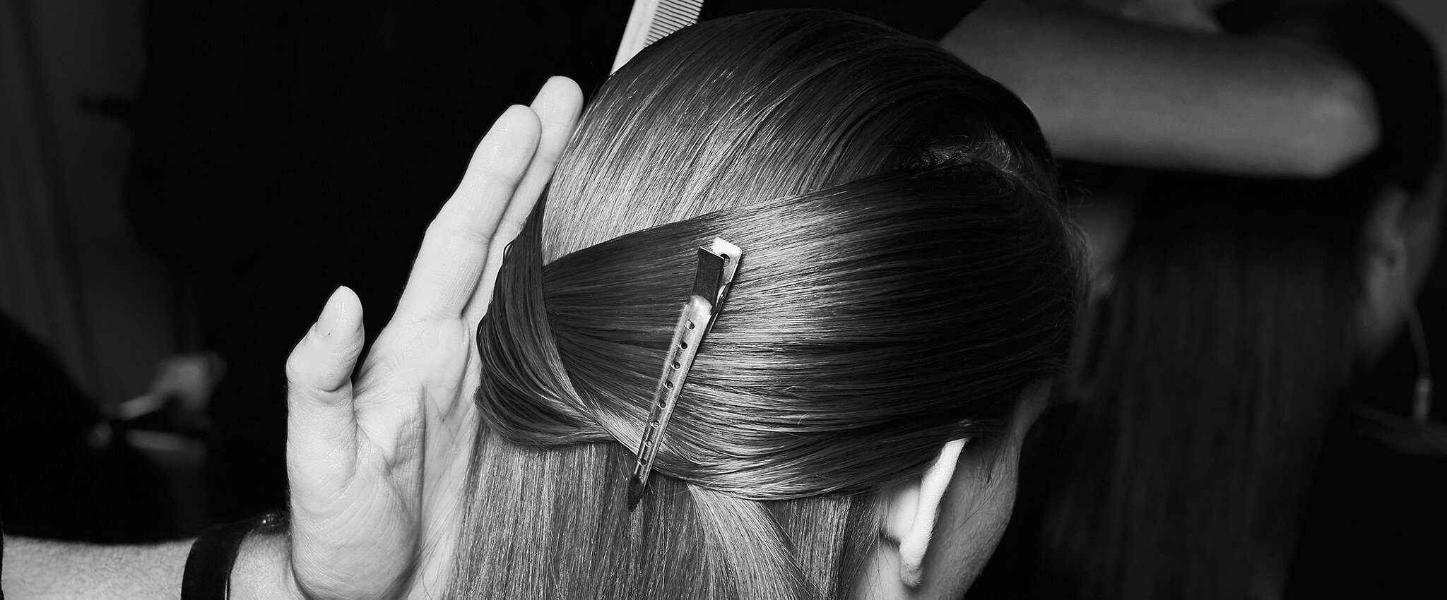 Fekete-fehér, közeli kép egy modell fejéről oldalnézetben, hajának egy része hajsütővasba fogva, a stylist keze éppen csak belóg.