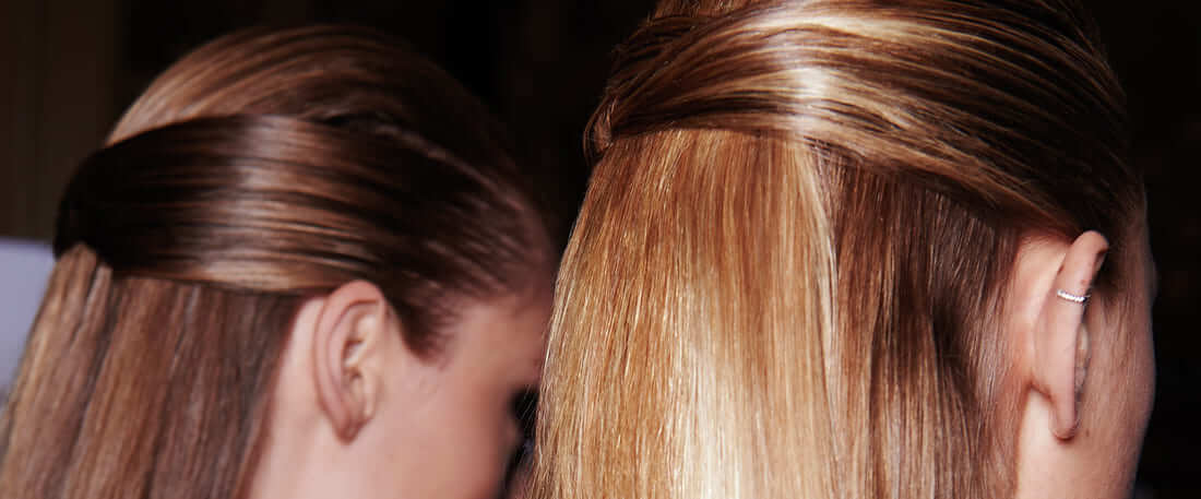 Close up of the side of two model's heads, with their hair in the same style, with the side sections wraped around the back of the head in a loose braid 