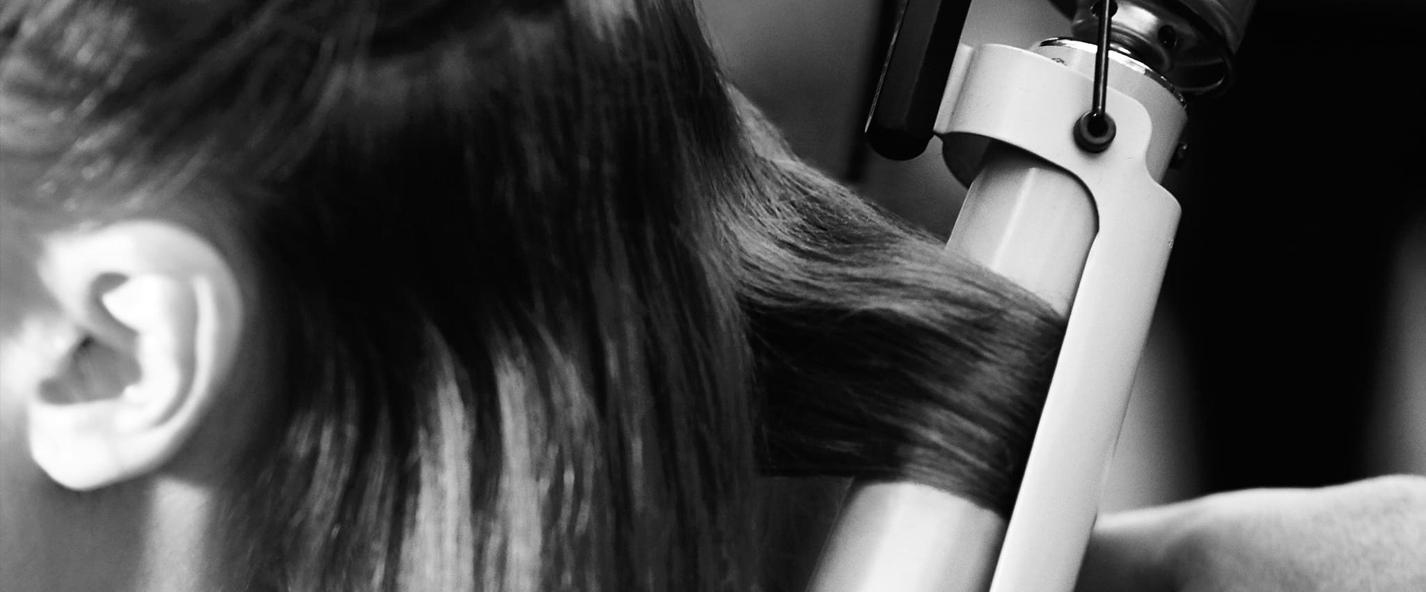 Black and white close up on the side of a model's head with a section of her hair in curling tongs and a stylist's hand just visible