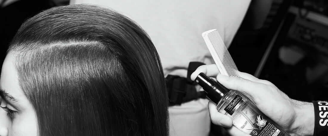 A stylist applies hairspray to a model's long dark, oily hair.