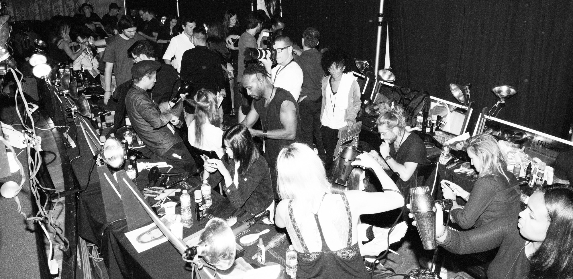 Two stylists braiding a model's hair with other models and hairdressers in the background 
