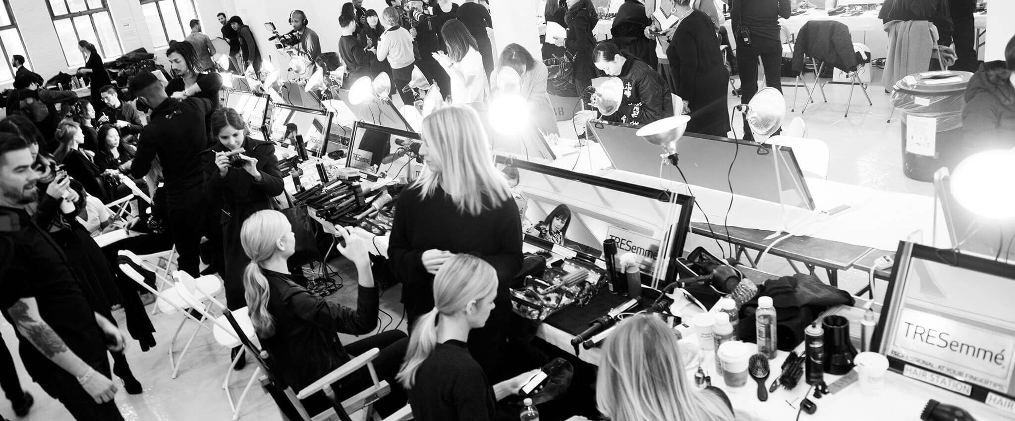 Models sitting in front of mirrors with hair stylists, photographers, styling tools and hair products 