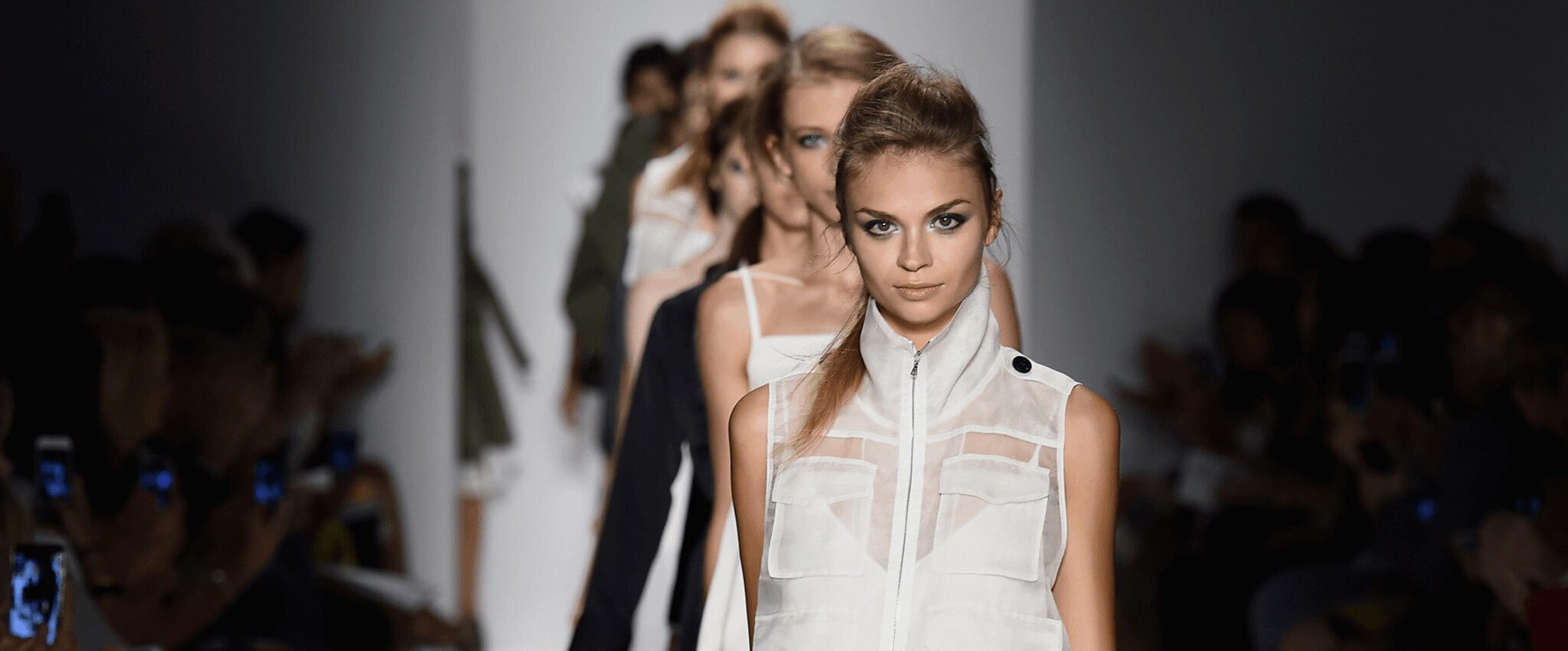 A line of models on the runway with a model wearing a white sleeveless jacket with pockets  