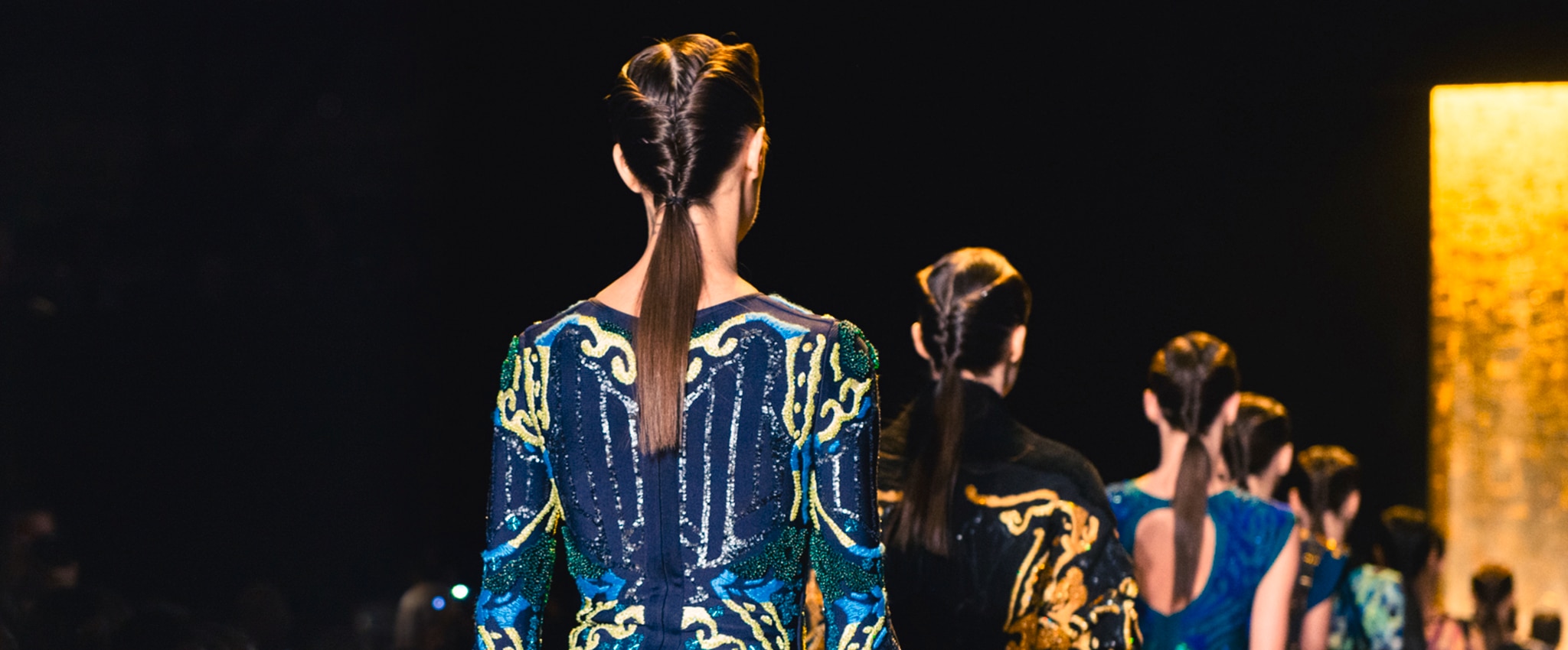 Photo of a models on the catwalk with their back to the camera, wearing colourful dresses with their hair in ponytails 