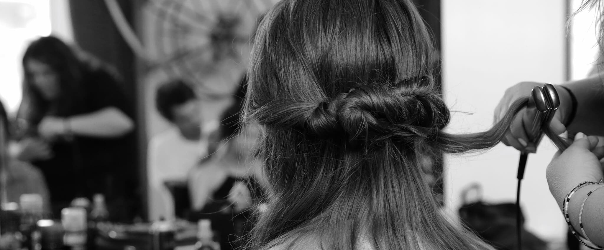Black and white backstage close up of the back of a model's head, with her long dark hair in a loose braid, with someone straightening a section of her hair with irons  