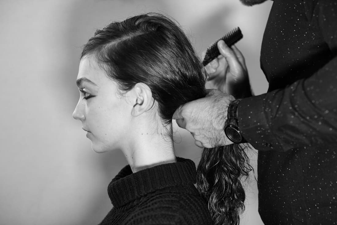 Black and white side shot of a model's head and shoulders with the hands and shirt of a stylist visible working through a section of her long dark hair with a long tail comb 