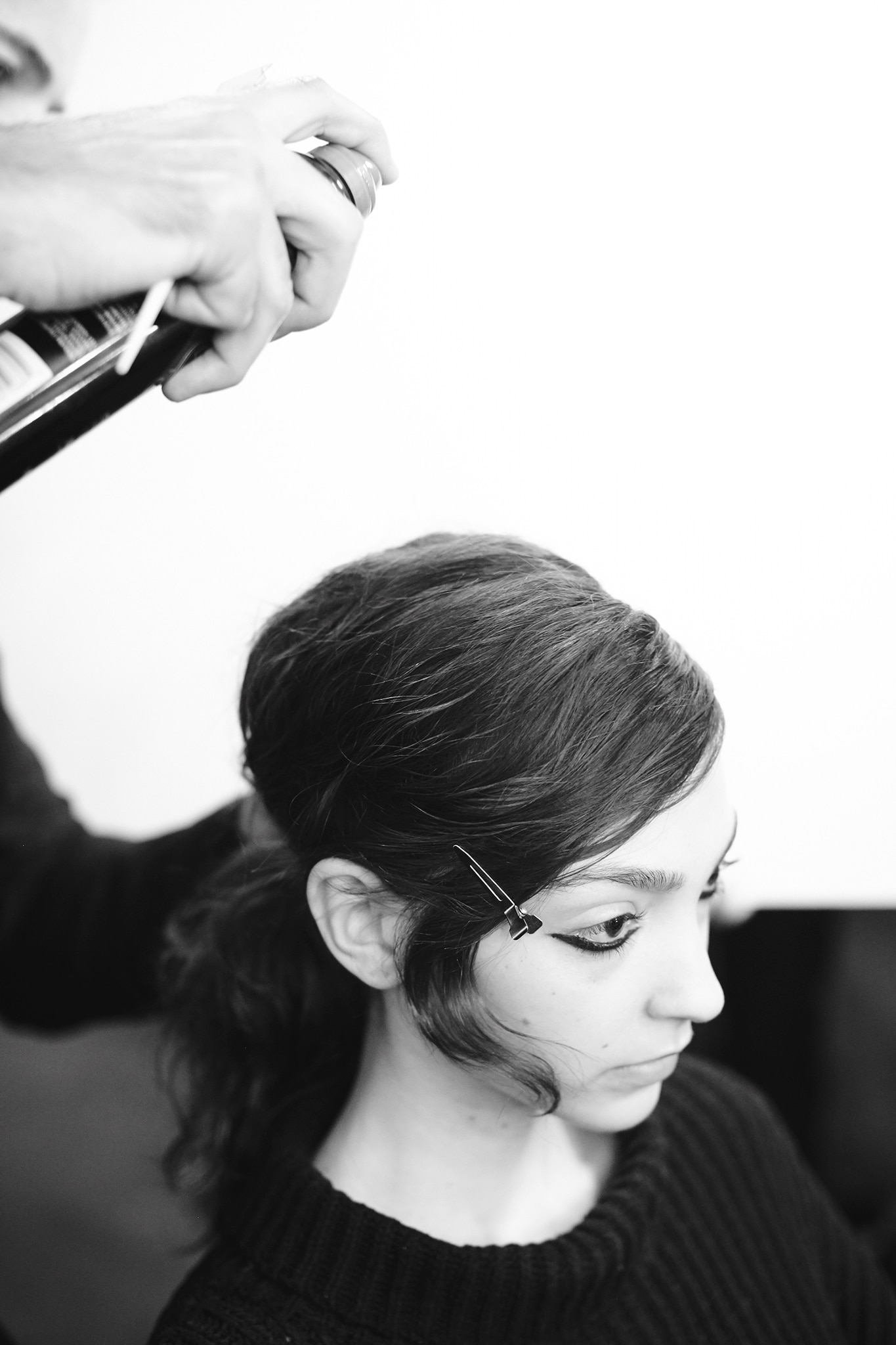 Uma modelo com cabelos escuros e compridos, com um rímel marcante nos olhos e um cabeleireiro aplicando spray para cabelo.  