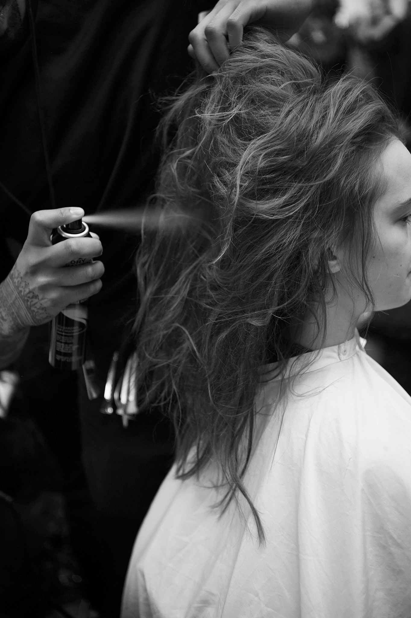 A model's head and shoulders with a stylist applying spray to her long curly hair