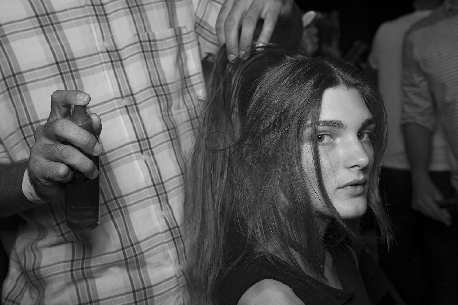 Una modelo con su largo pelo castaño, húmedo, a quien un estilista le aplica spray con un producto fuera de la foto