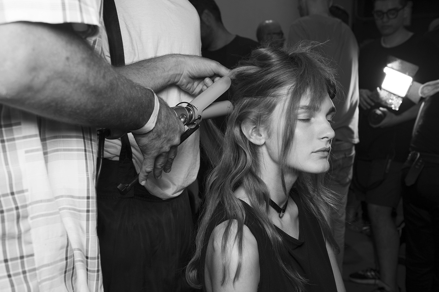 Un estilista aplicando un rizador a un mechón del pelo de una modelo con un fotógrafo entre bambalinas