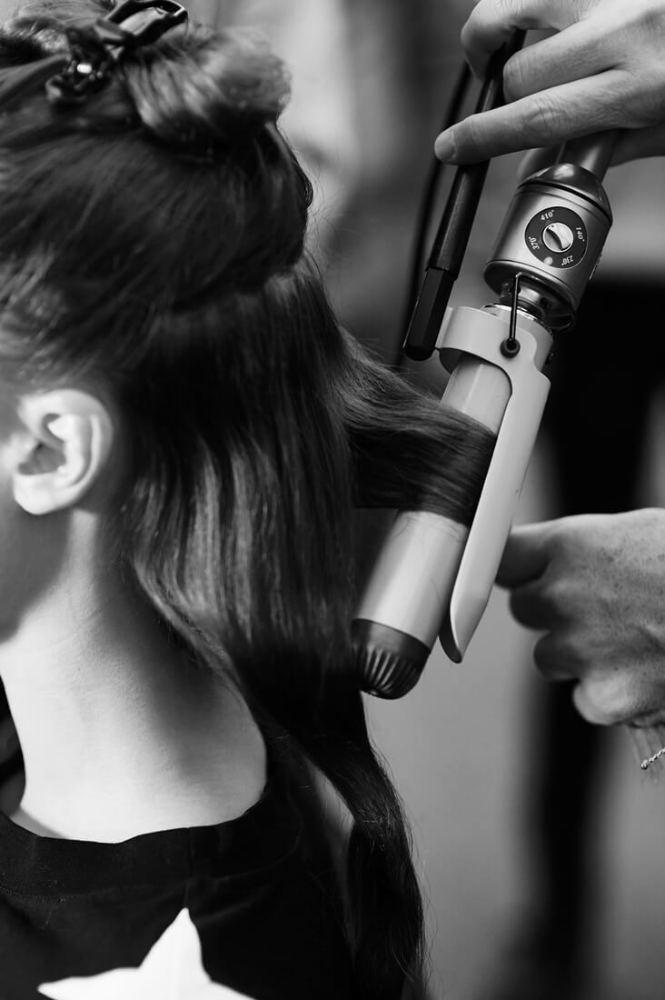 A model with her hair up in a clasp and a stylist applying a curling wand to a section of it 