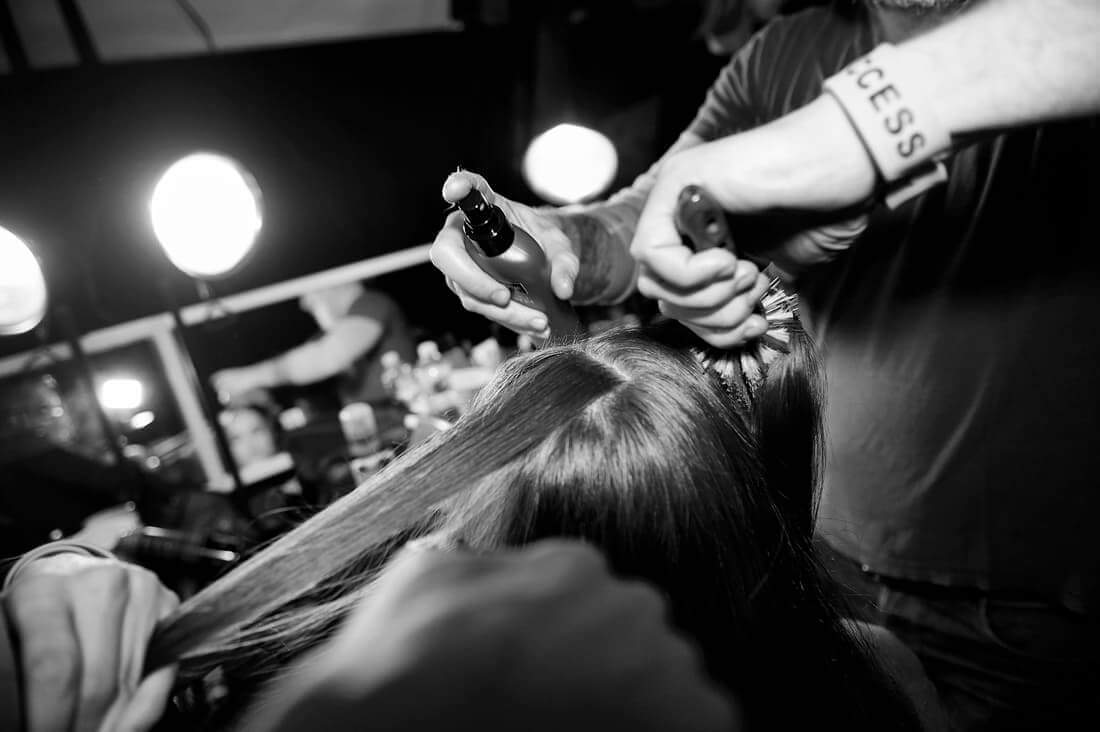 Two stylists working on a model's hair backstage with brushes and styling products.