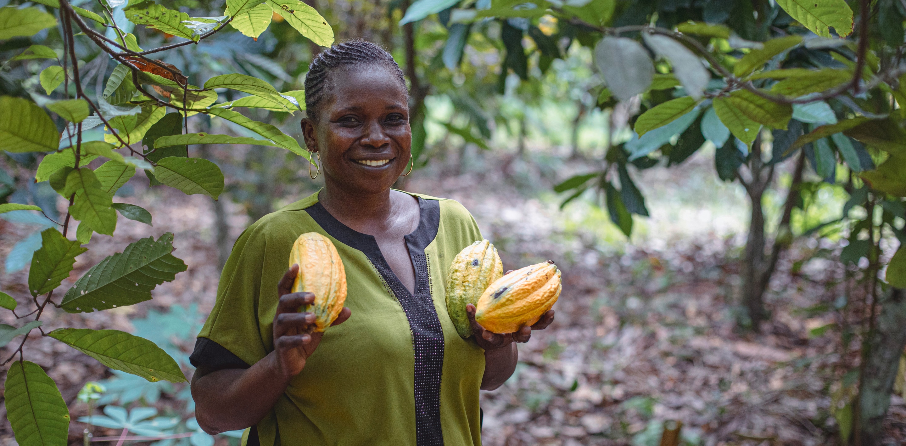 Granjera de cacao sonriente con un top verde y sosteniendo tres vainas de cacao amarillas.s
