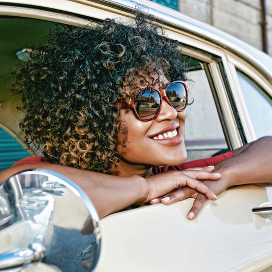 Mujer con cabello enrulado sonriendo, con anteojos de sol, asomanda por la ventanilla del auto. 