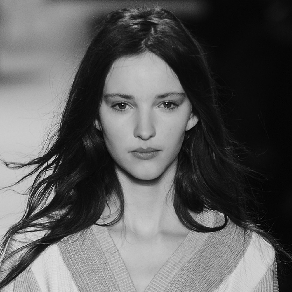 A model on the runway, with long dark brown hair, wearing a grey and white V-neck top