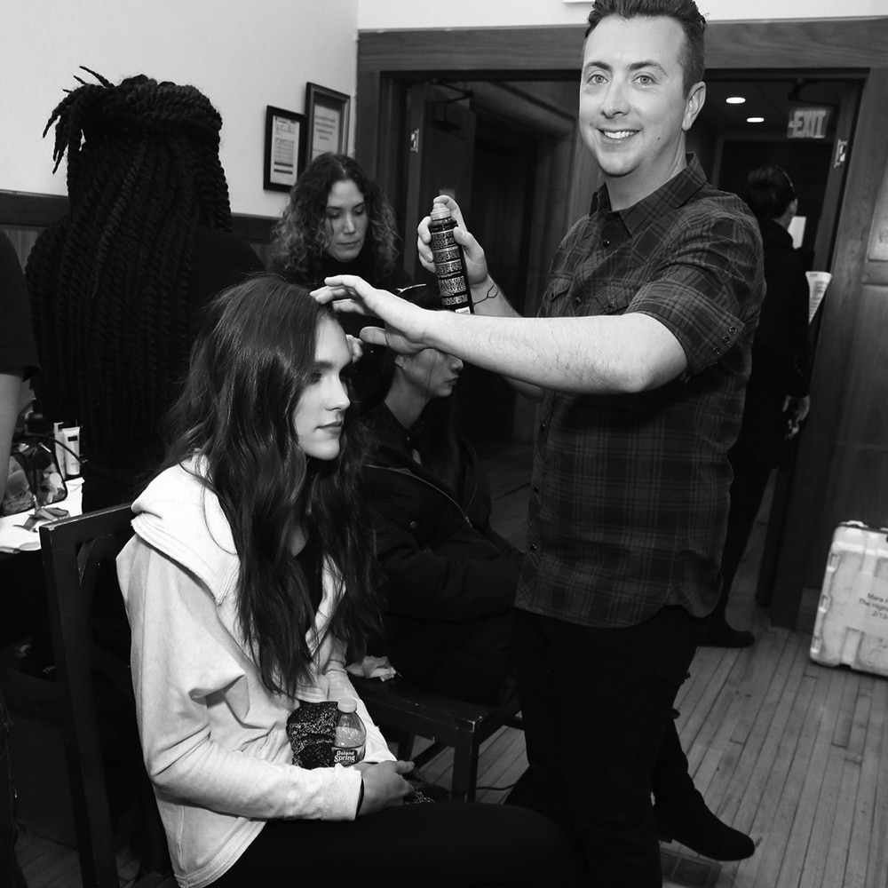Stylist Tyler Laswell finishing a model's hair with spray, with models and stylists in the background